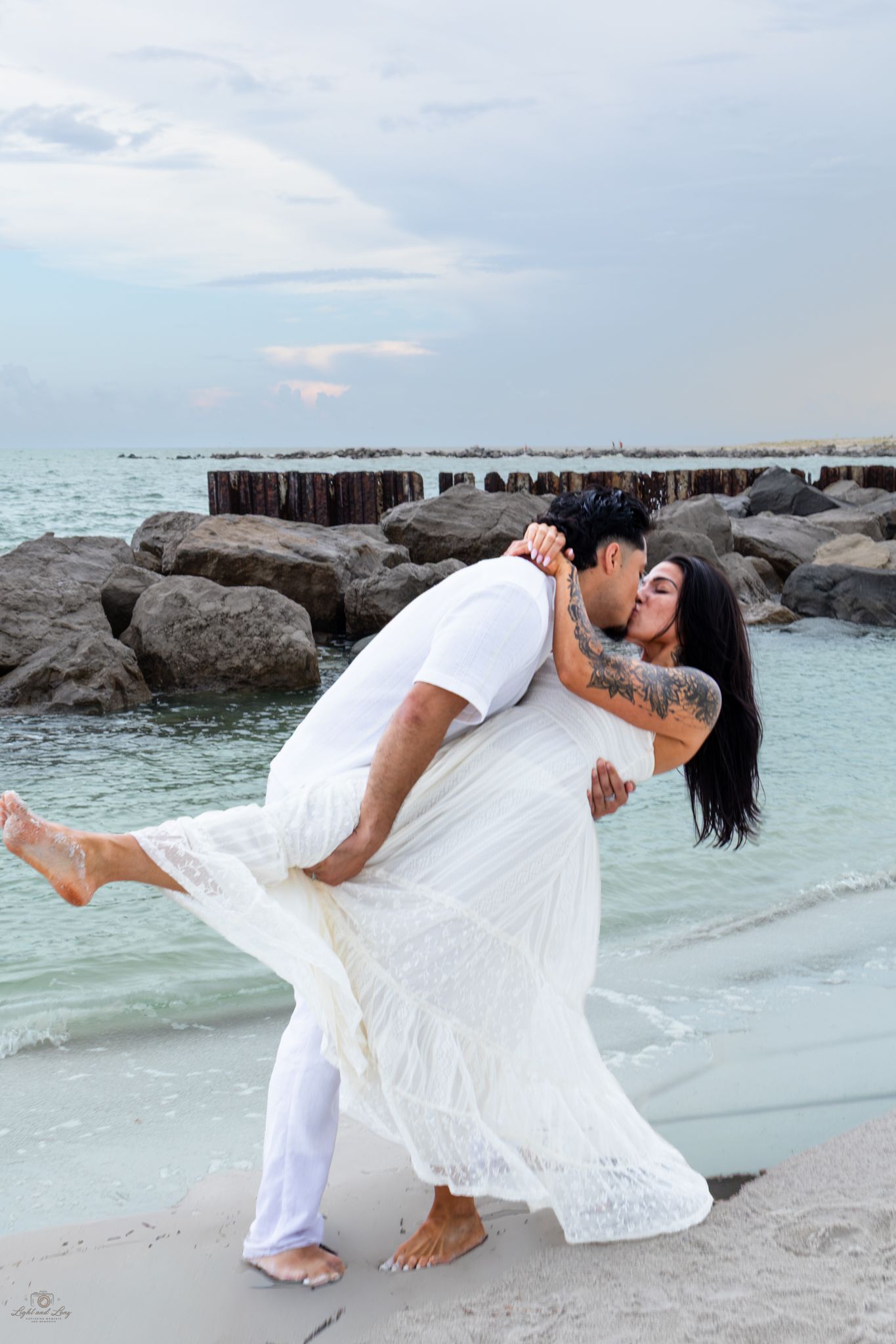 Couple sharing a romantic dip kiss on a sandy beach, dressed in white, with turquoise ocean waves and rocks behind them.