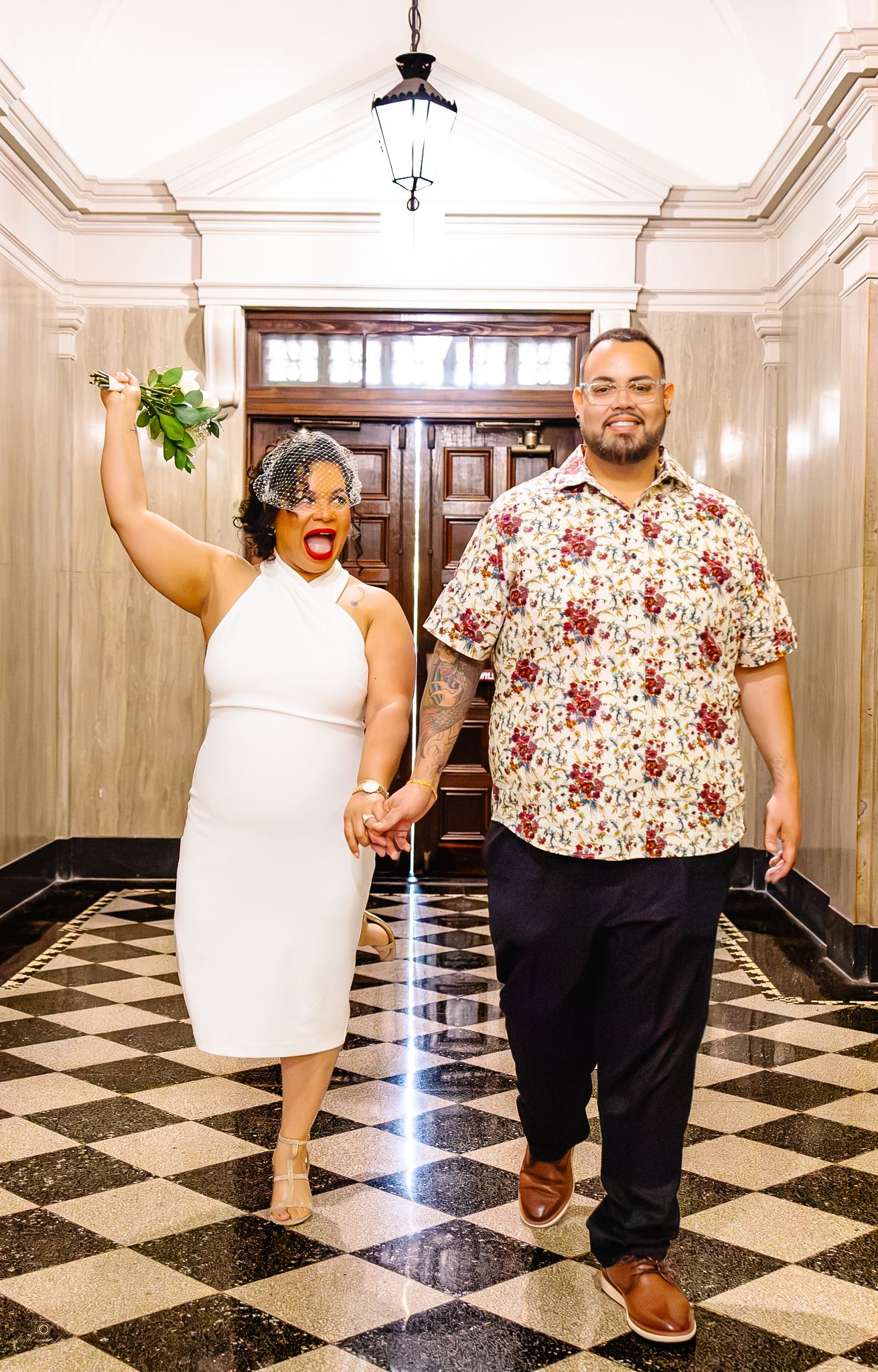 Joyful bride in a white halter dress raises her bouquet and cheers while holding hands with her groom on a black and white tiled floor.