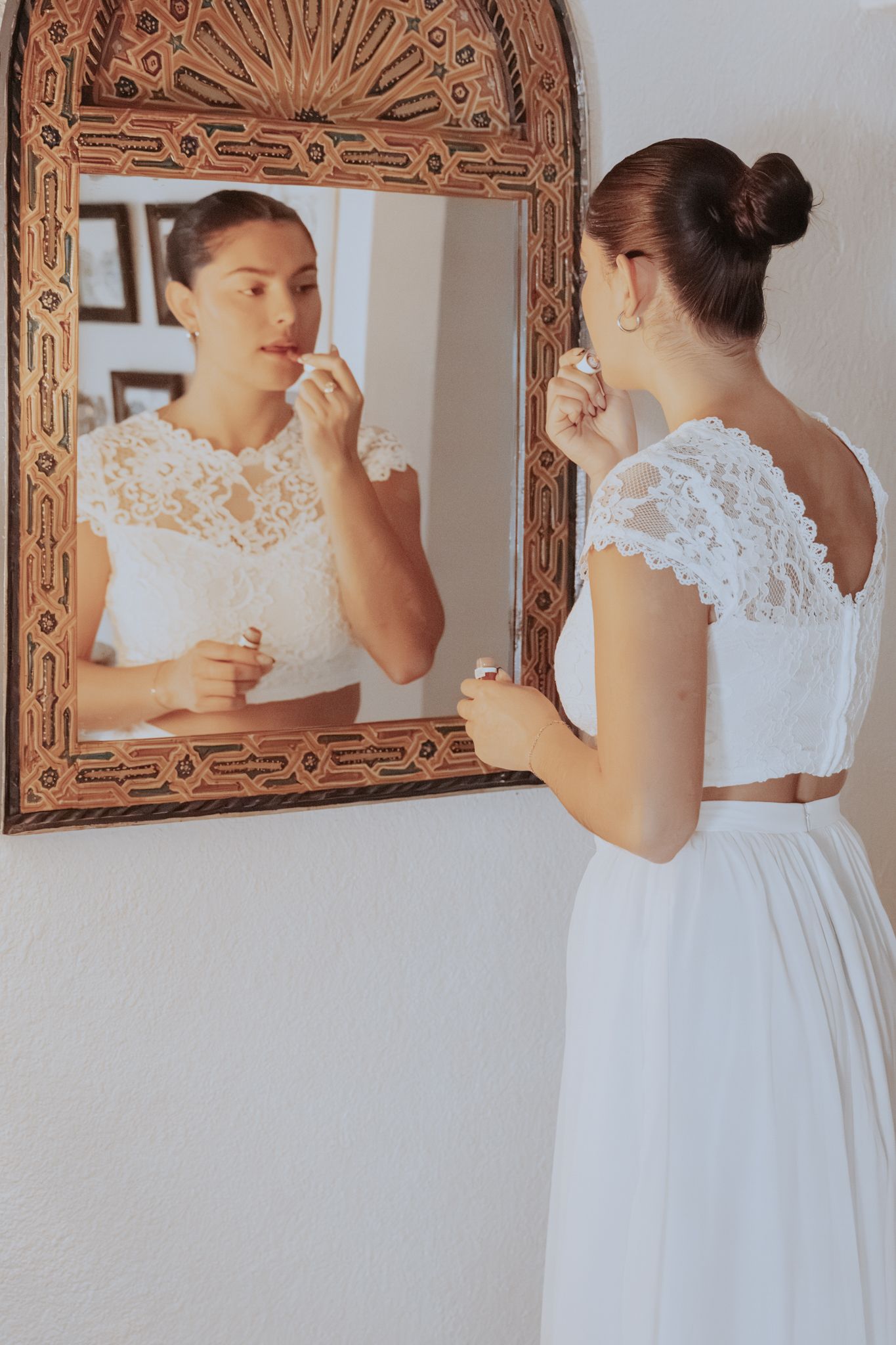 Woman in a white lace top and flowing white skirt applies lipstick while looking into an ornate carved wooden mirror.