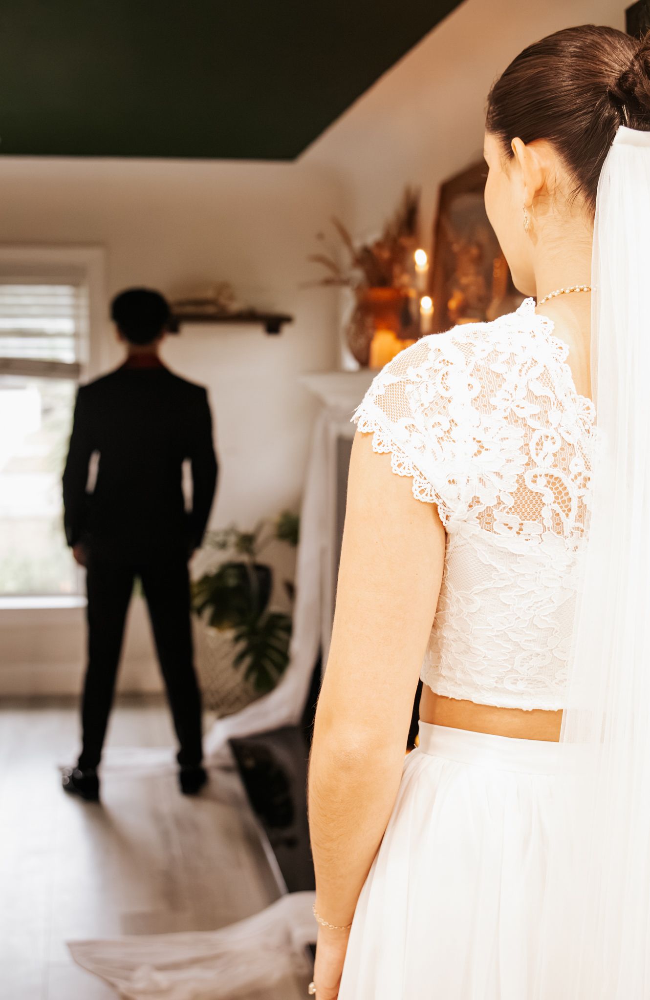 Bride in a lace wedding top and veil faces a groom in a black suit standing with his back turned in a bright room.