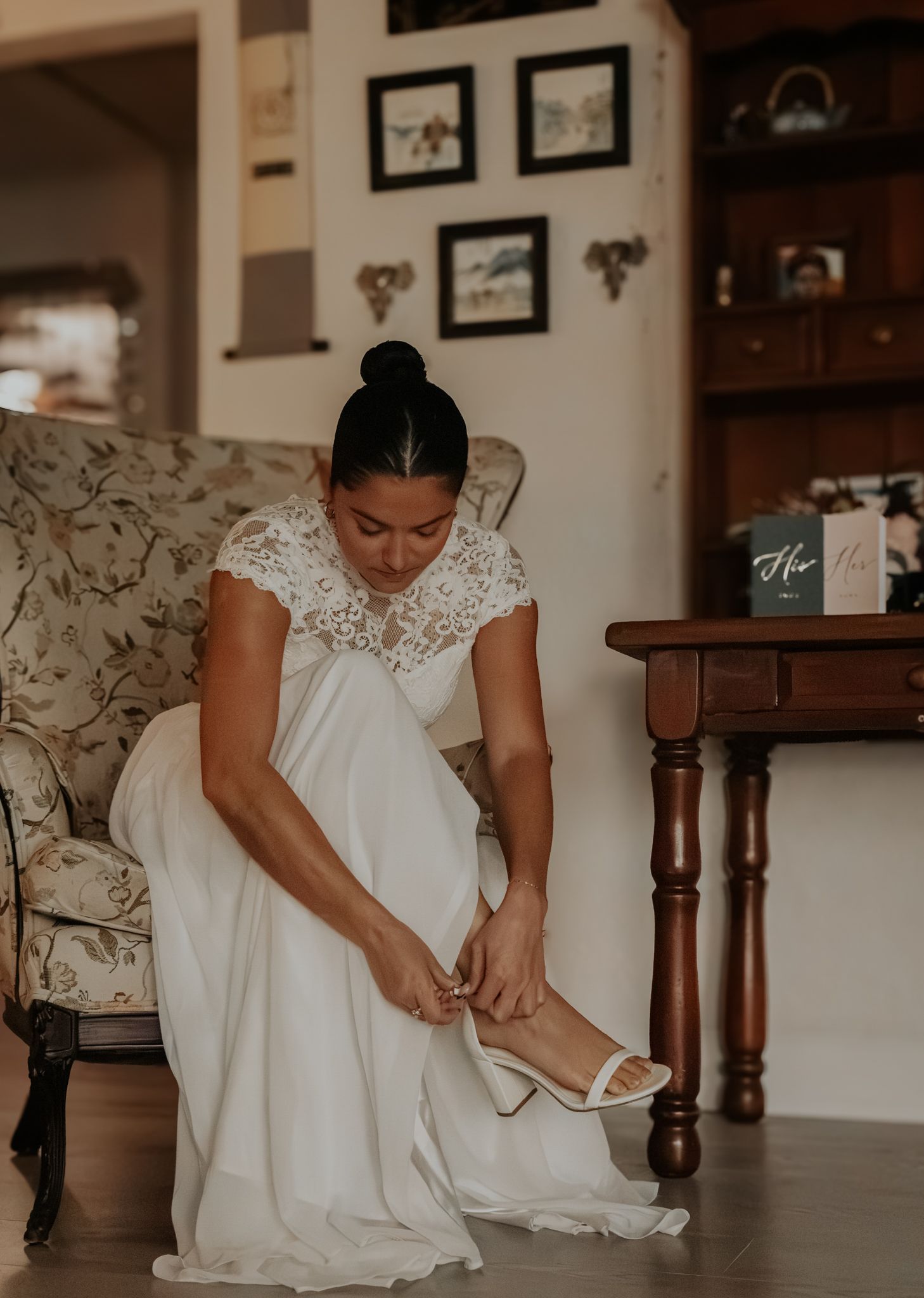 Bride in a white lace wedding dress and hair bun sits on a floral armchair, fastening her white heeled sandal in a cozy, vintage-style room.