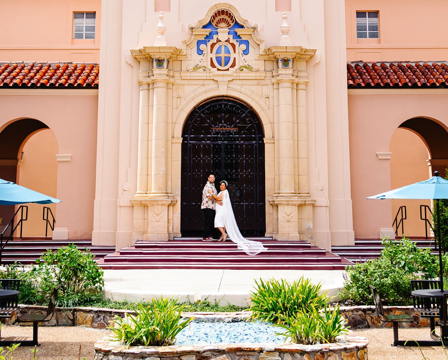 Newlyweds embracing on steps of a Spanish-style pink building with ornate arched entrance doors.