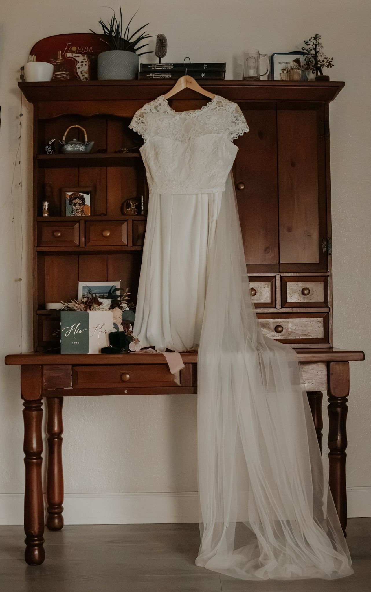 A white lace-bodice wedding dress with a long tulle train hangs on a wooden hutch desk decorated with framed photos, flowers and a teapot.