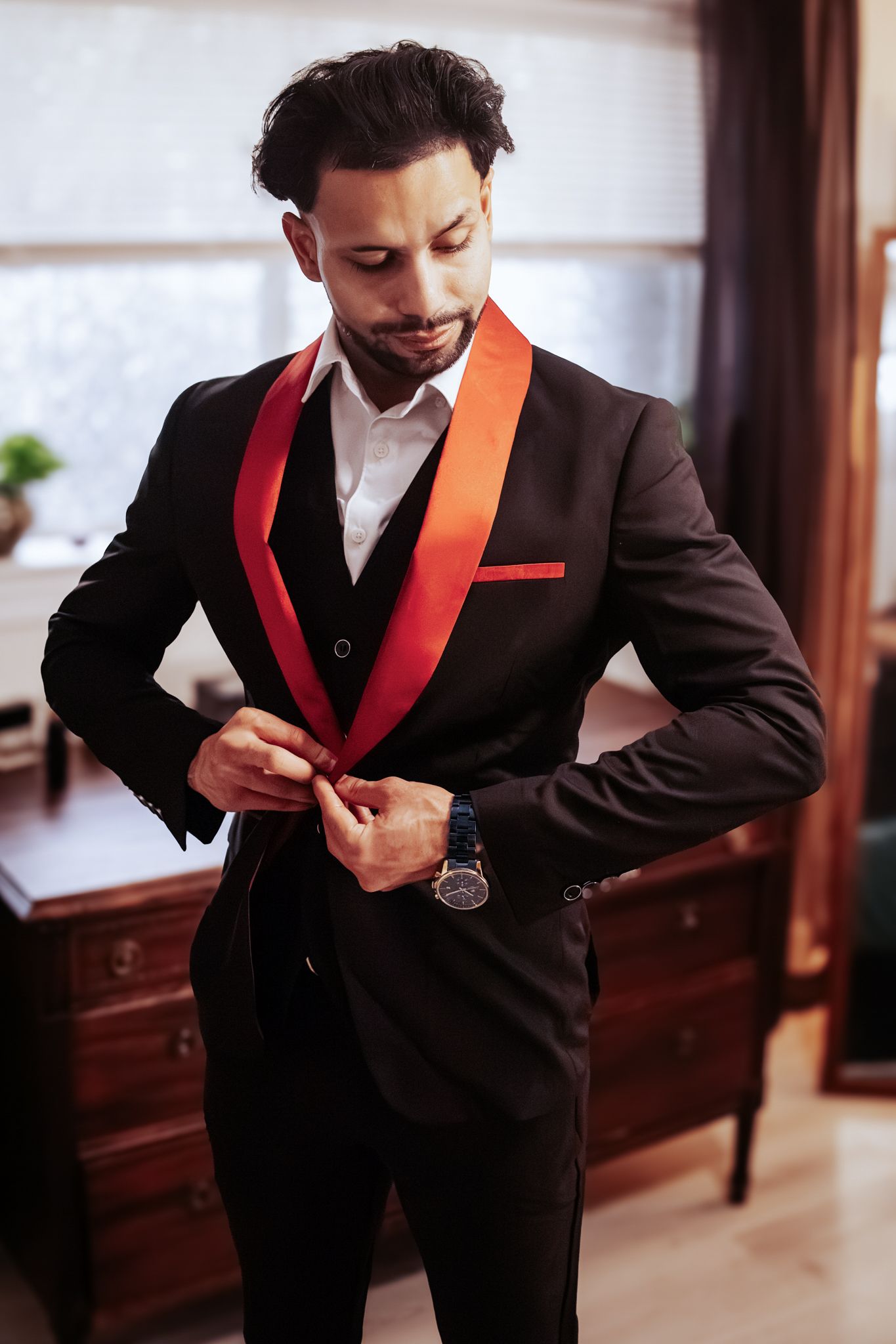 Groom in a black tuxedo with red satin shawl lapels buttoning his jacket beside a wooden dresser while getting ready.