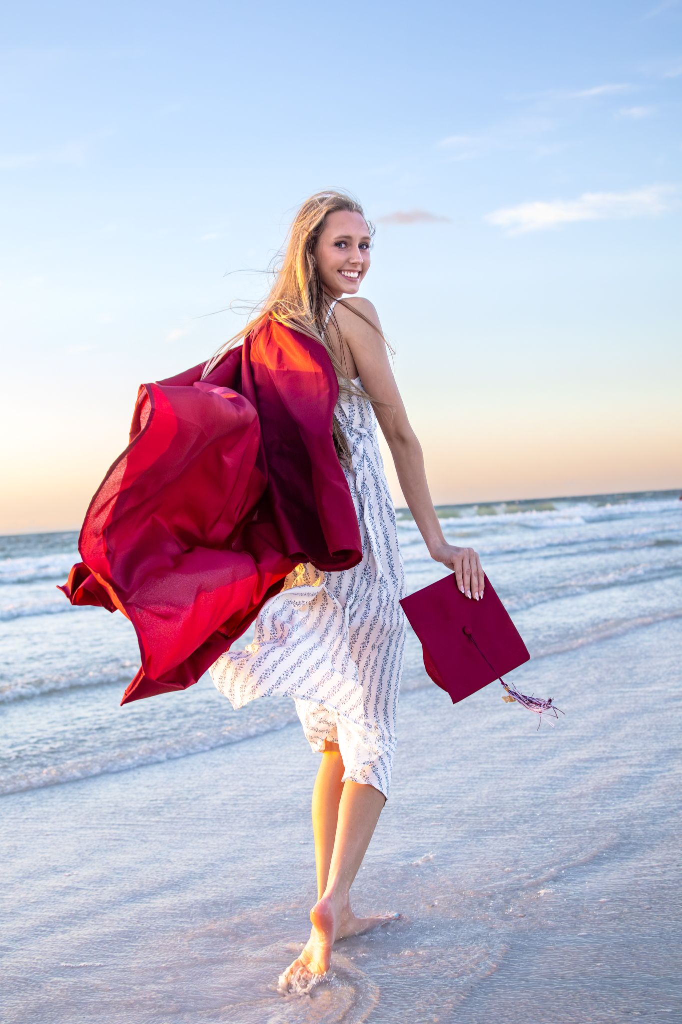 Smiling graduate walks barefoot on beach at sunset, holding red cap and gown flowing in the wind.