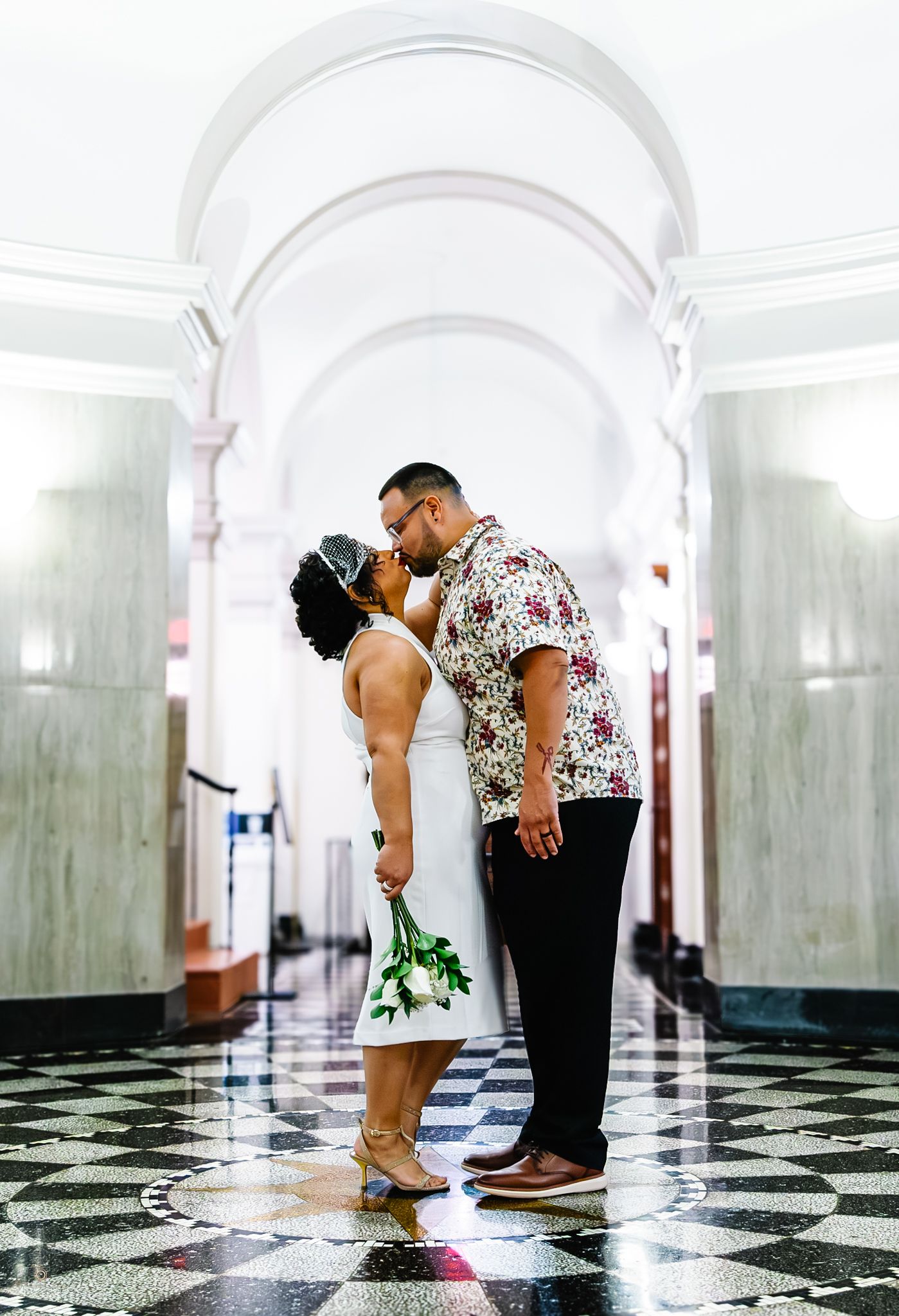Wedding couple kissing in a grand marble corridor with arched ceilings and checkered tile floors.