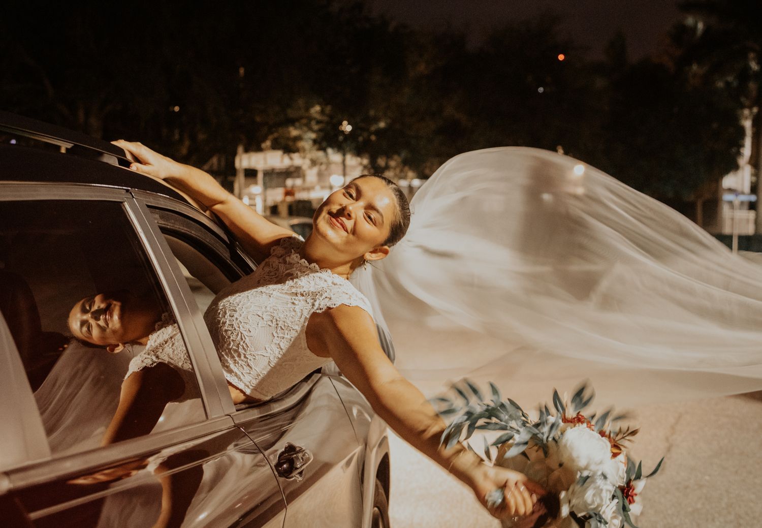 Smiling bride in a white lace dress leans out of a car window at night, her long veil flowing behind her as she holds a floral bouquet.