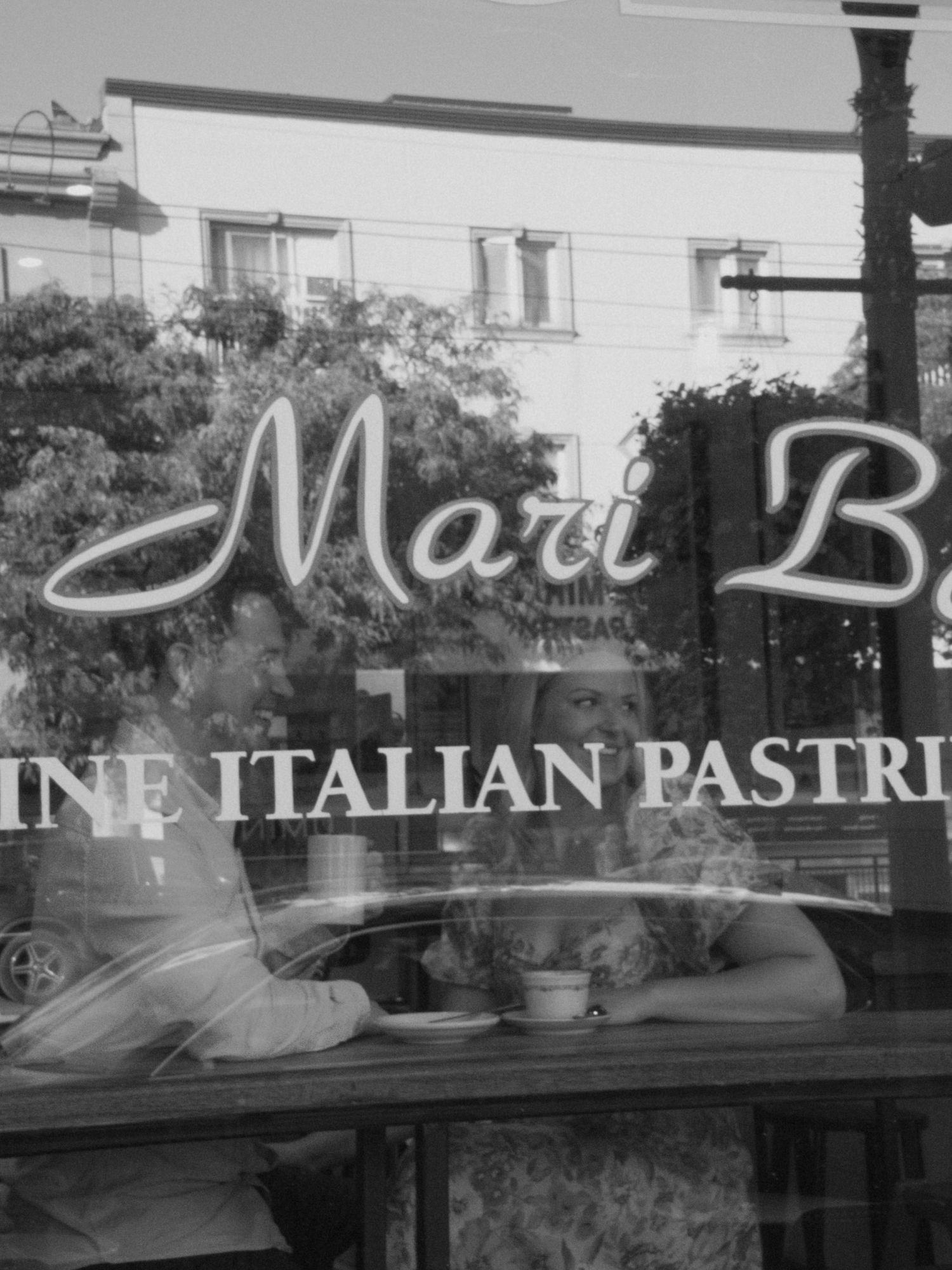 Black and white image of a couple seated inside a café, seen through a window with reflections and signage