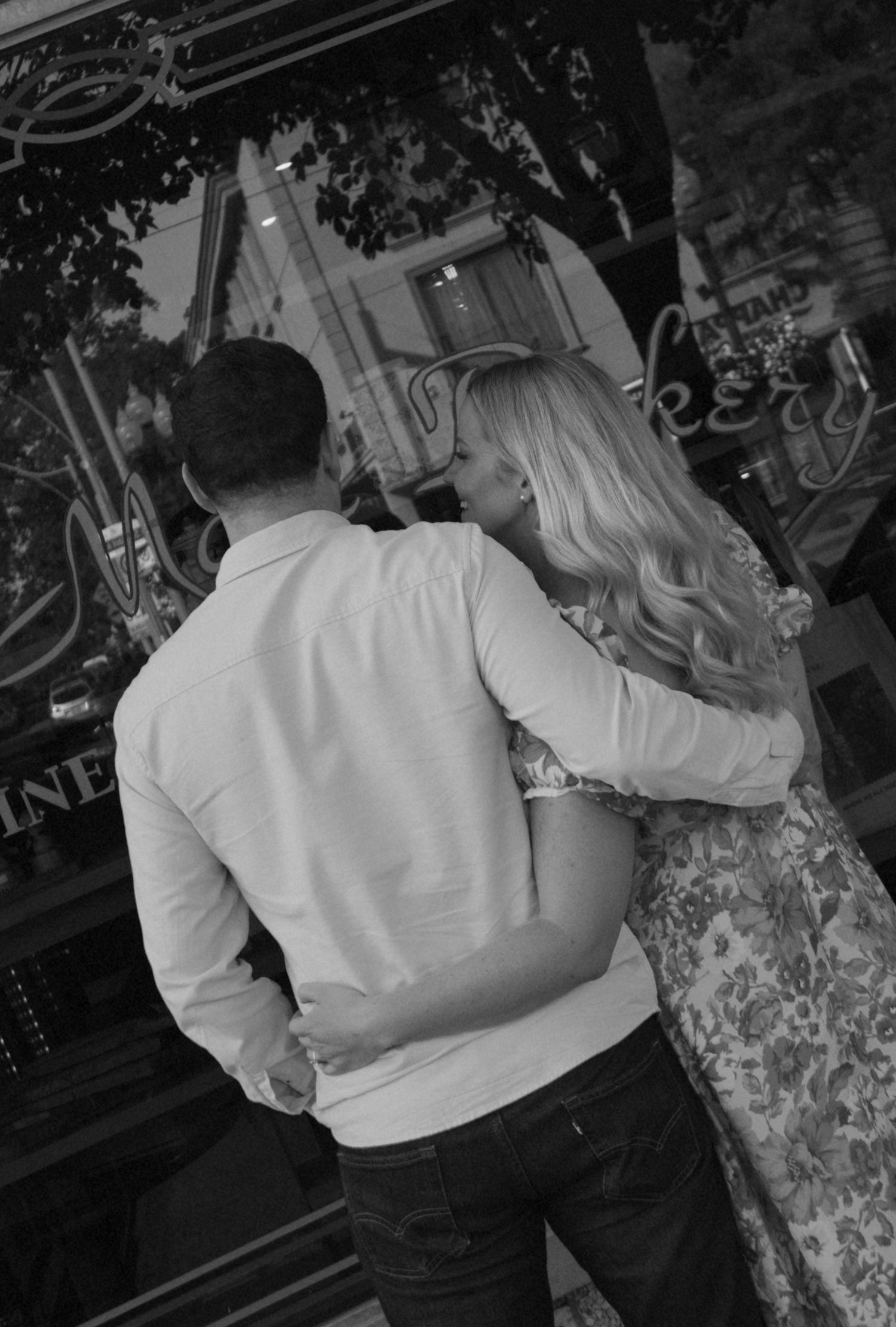 Couple embracing from behind outside a storefront, captured in a candid black and white moment