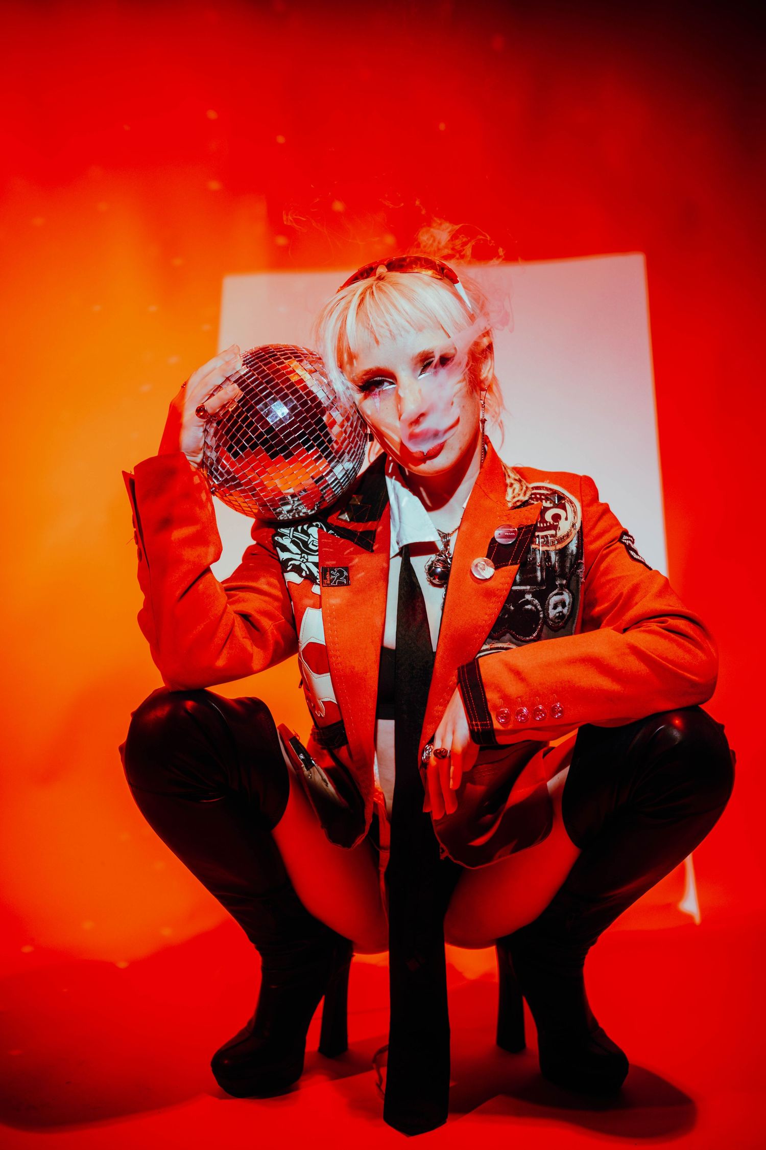 Blonde person in red jacket squats holding disco ball in vibrant red studio.