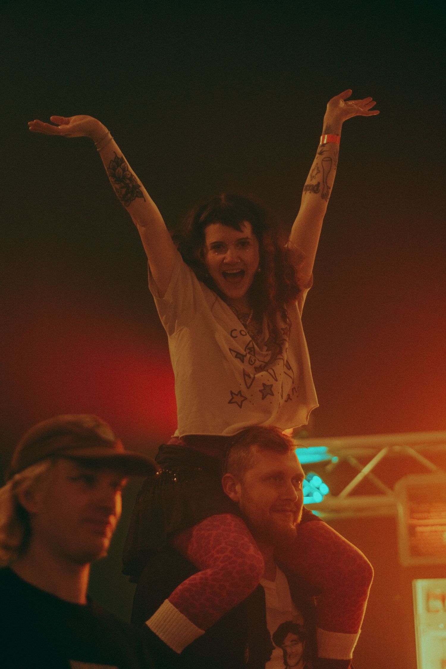 Excited woman with tattoos raising arms on mans shoulders at a concert.