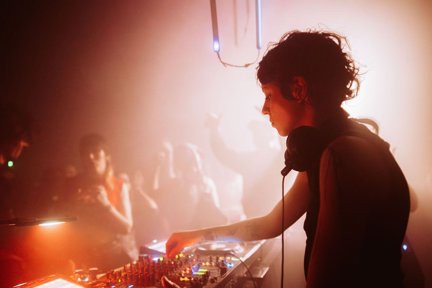 DJ mixing at turntables with headphones around neck in warm orange-lit nightclub.