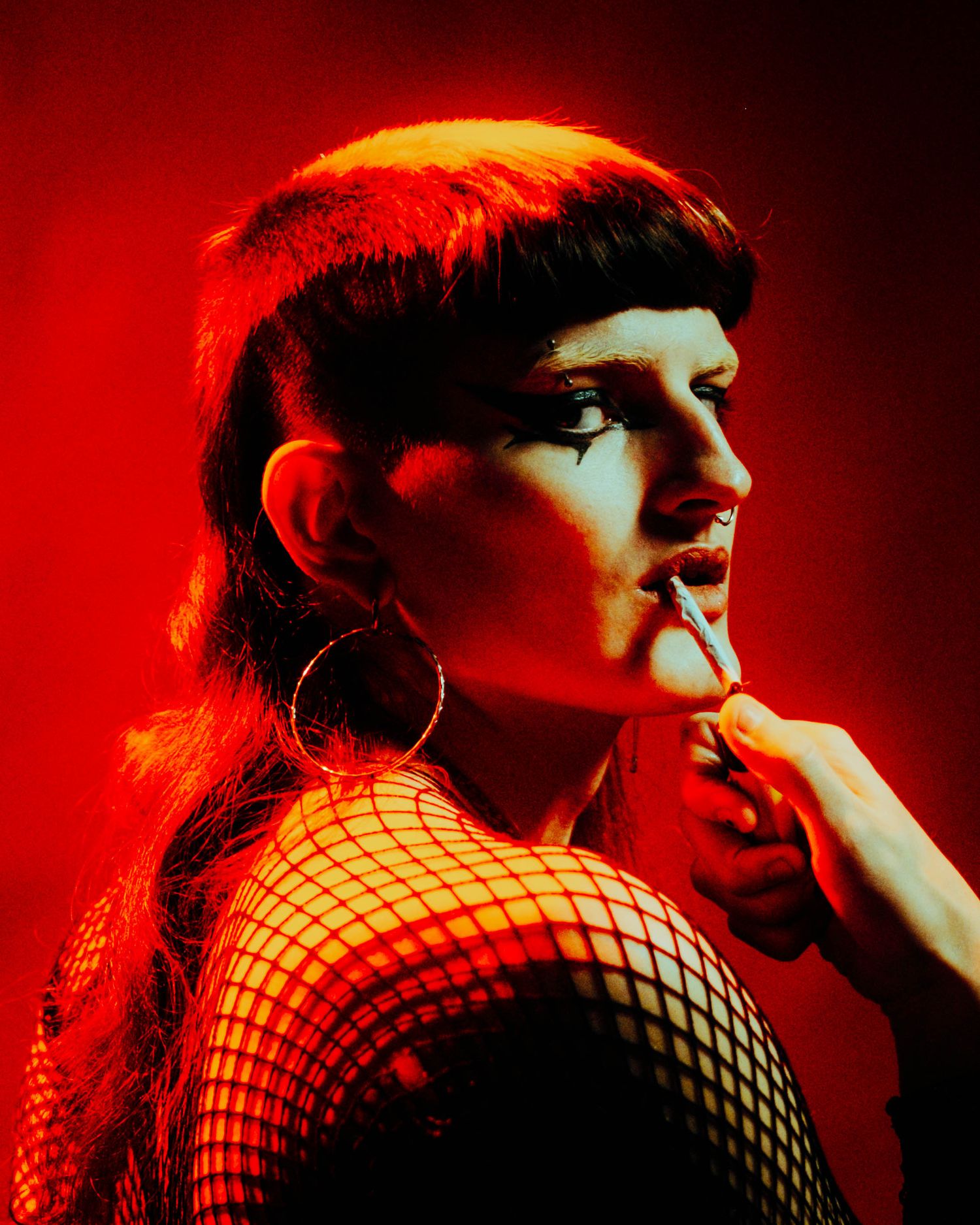 Person with bold makeup in fishnet top poses under red dramatic studio lighting.
