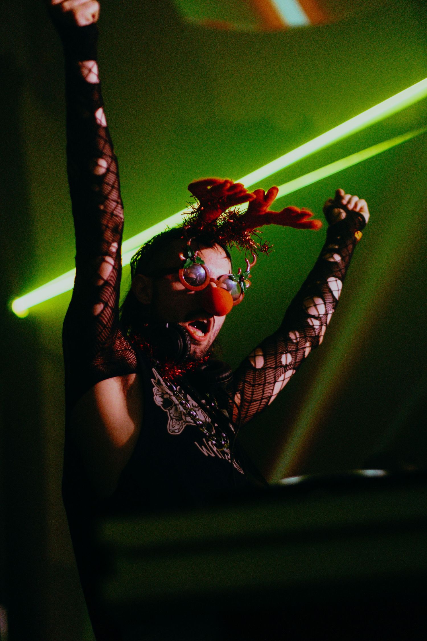 Energetic performer in fishnet outfit and clown nose dancing under green laser lights at a nightclub.