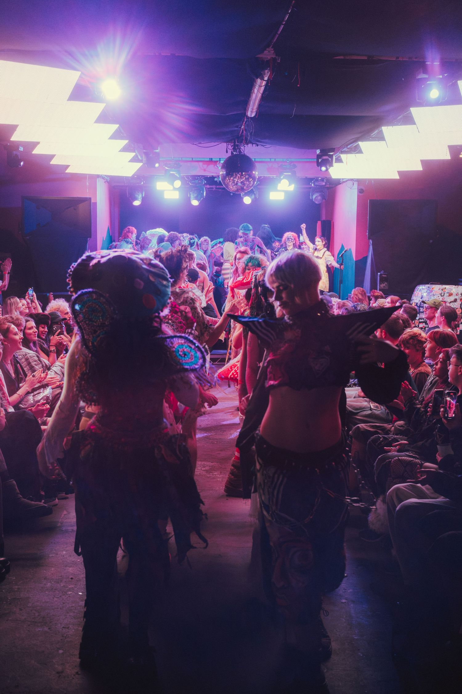 Two elaborately costumed performers walk through a cheering crowd toward a colorfully lit stage.