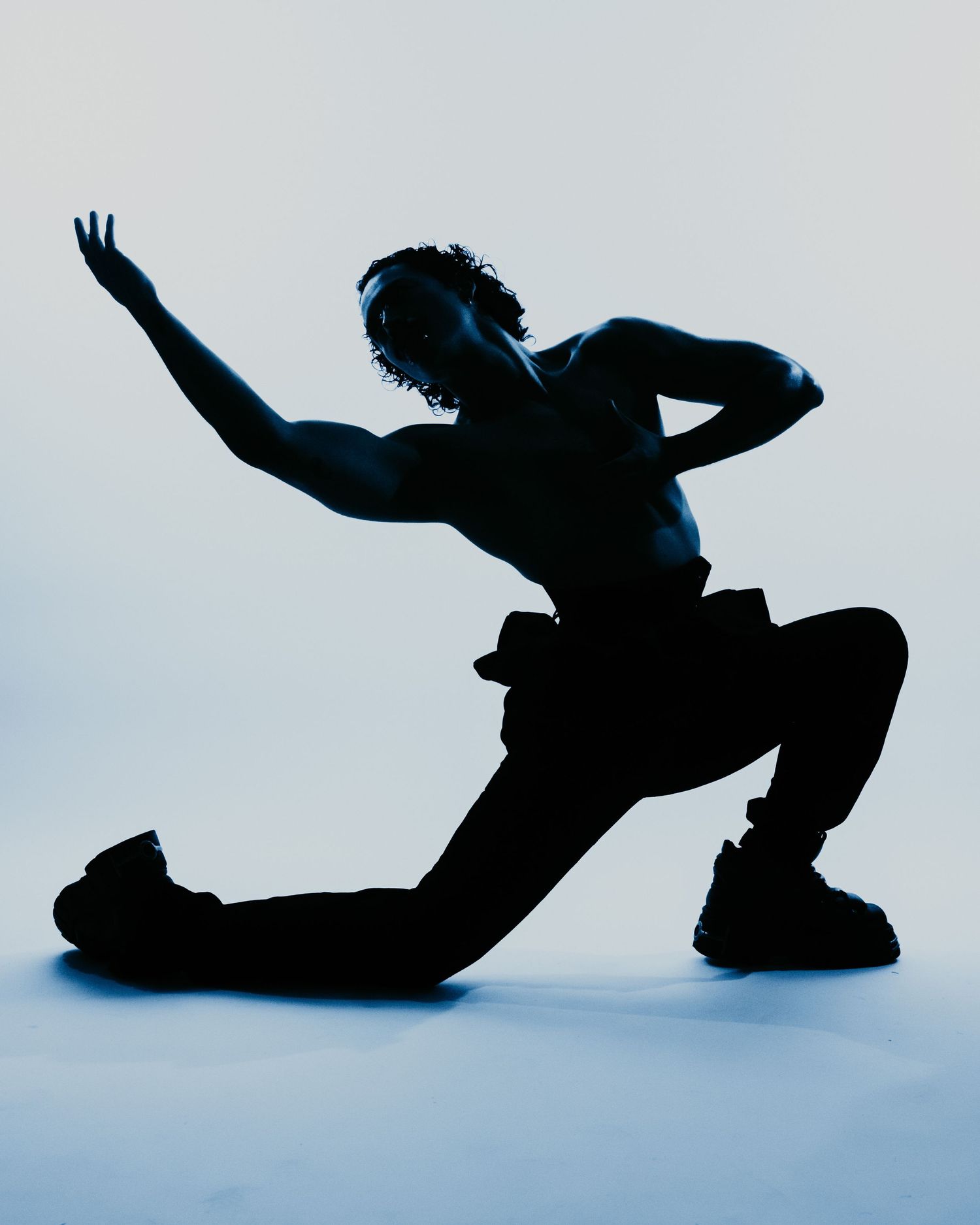Silhouette of shirtless dancer in wide lunge pose against light blue background.
