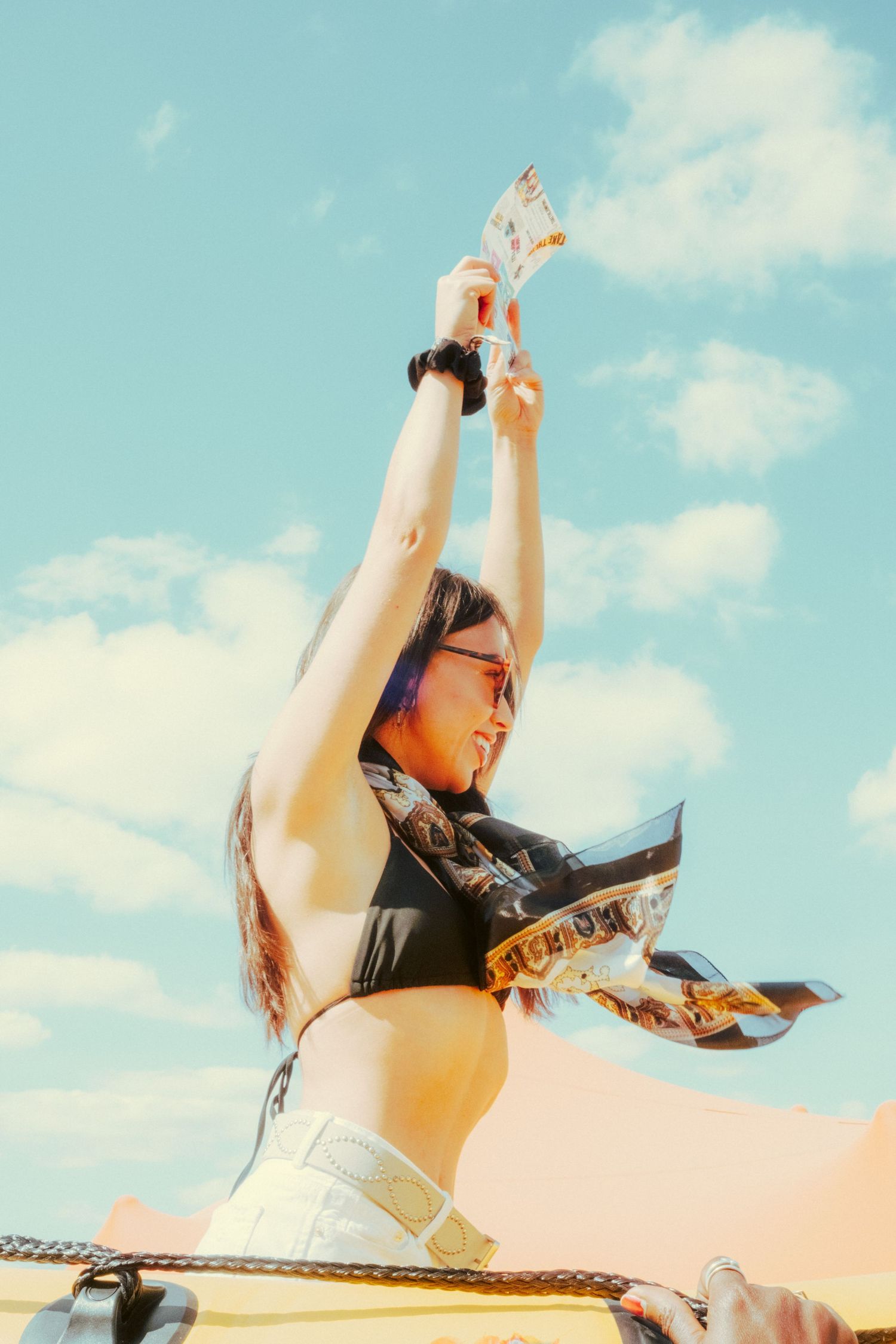 Young woman in crop top raising hands with money at an outdoor festival under blue sky.