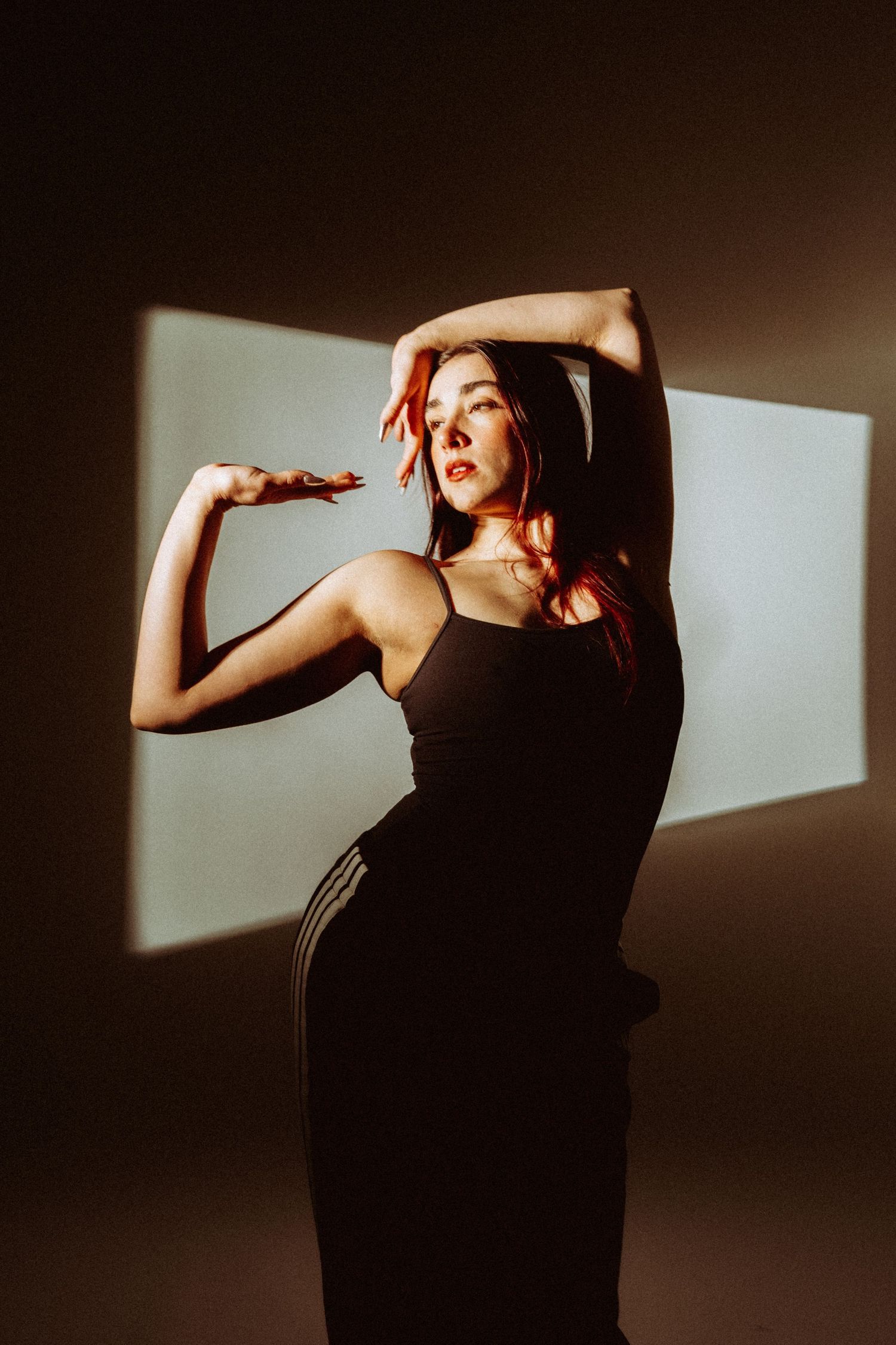 Woman in black dress poses gracefully with arm raised against shadowed wall.