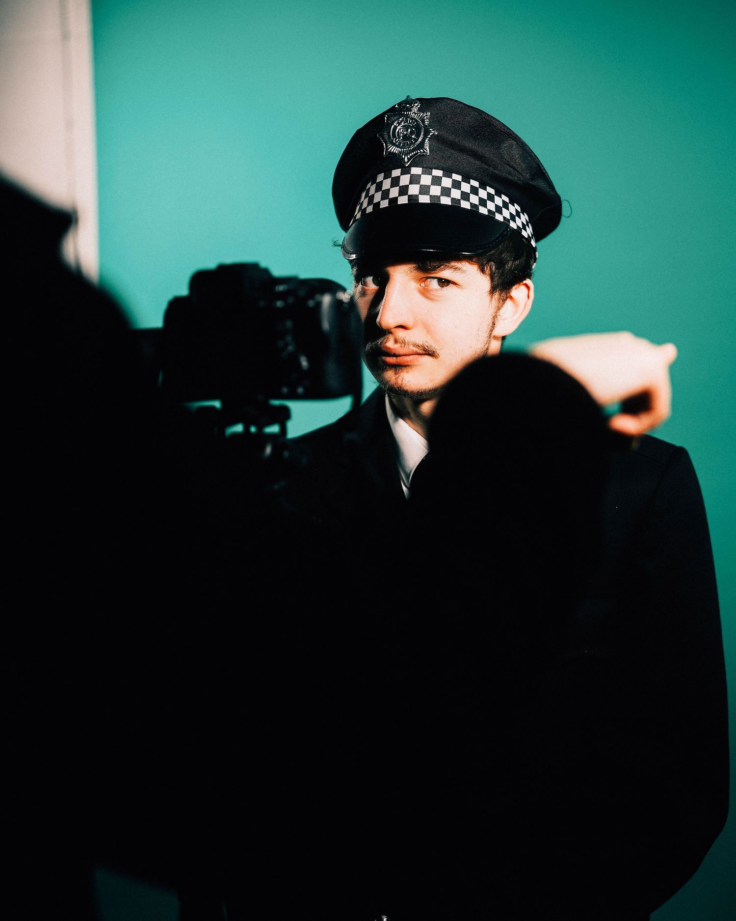 A young man in a police officer costume hat looks into a camera against a teal background.