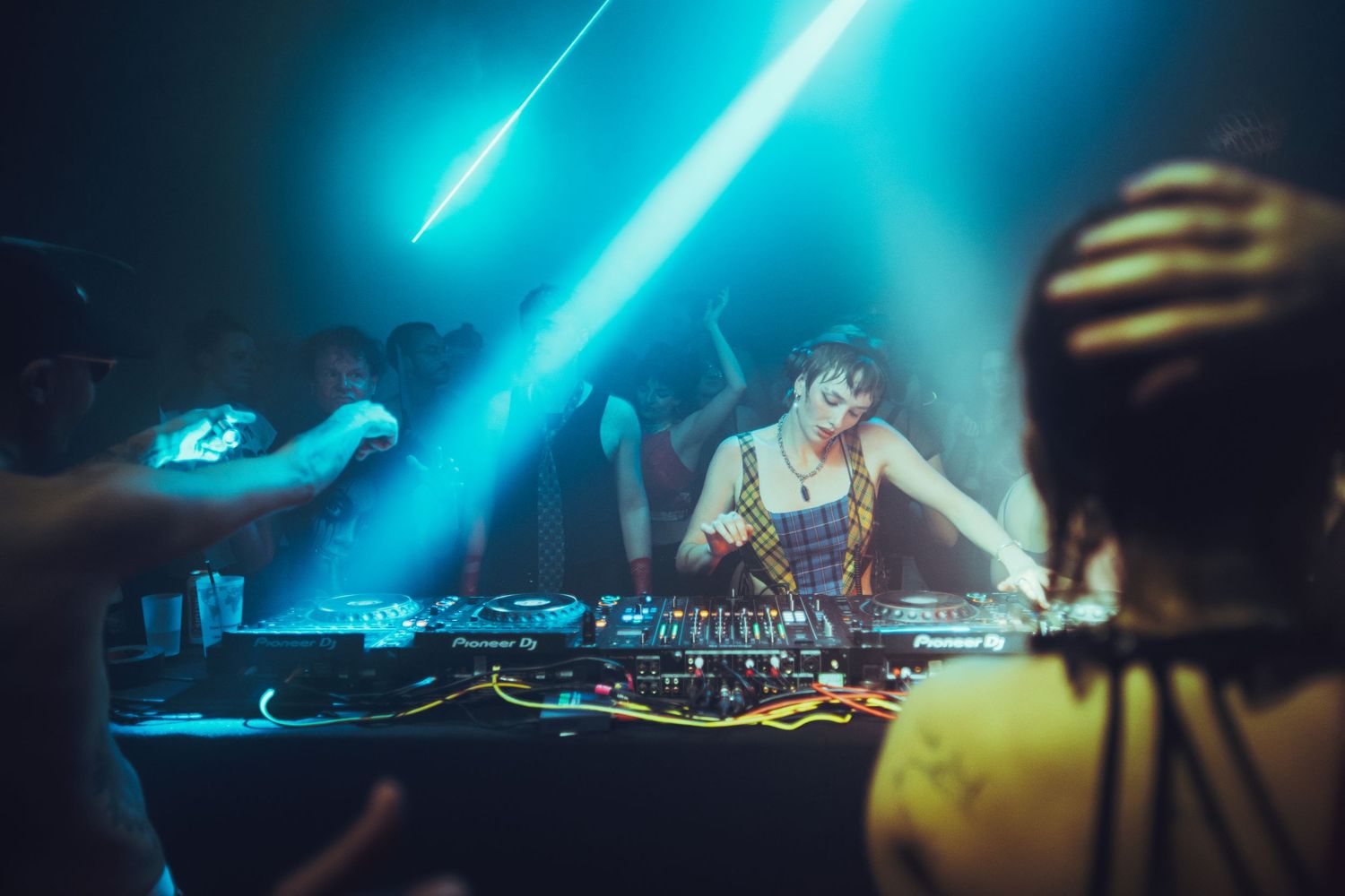 Female DJ performing at turntables under dramatic blue laser lights in a nightclub.