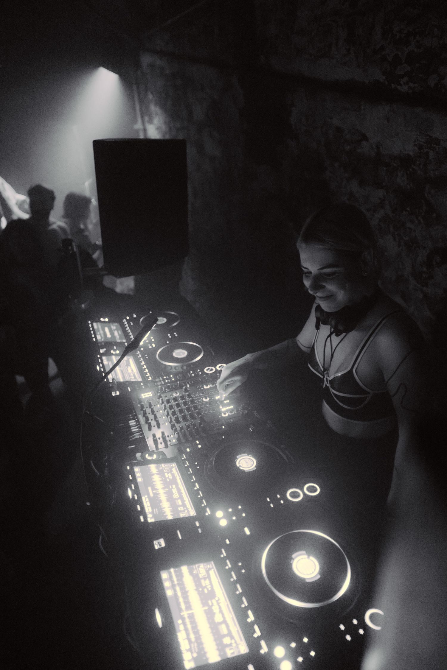 Female DJ operating Pioneer DJ decks in a dimly lit venue, captured in black and white.
