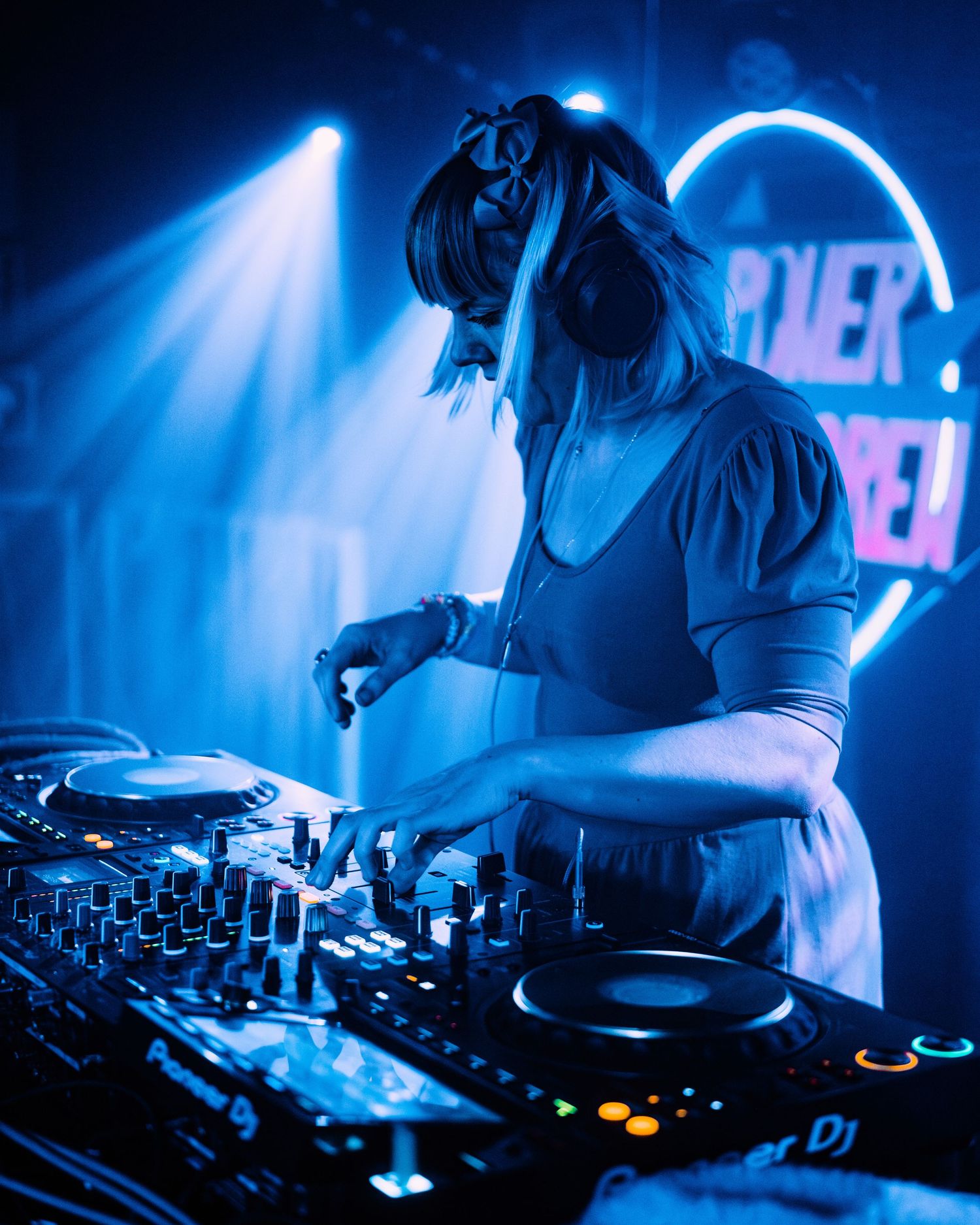 Female DJ wearing headphones performs at a professional DJ booth under dramatic blue stage lighting.