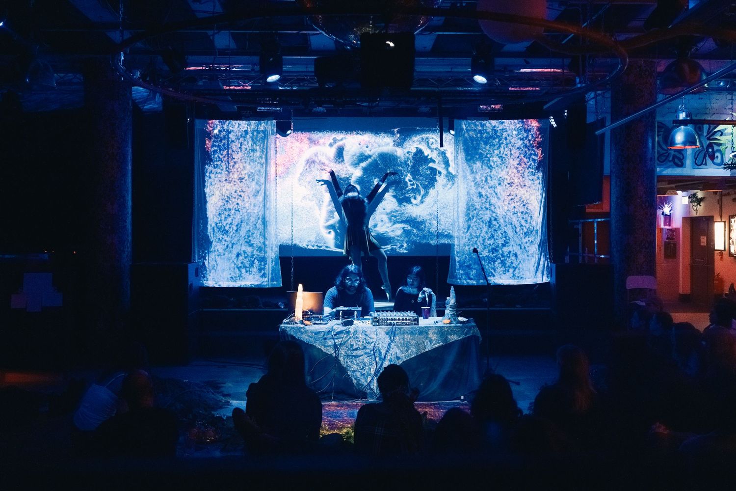 DJ performing under blue light with large projection screen at dark indoor venue.