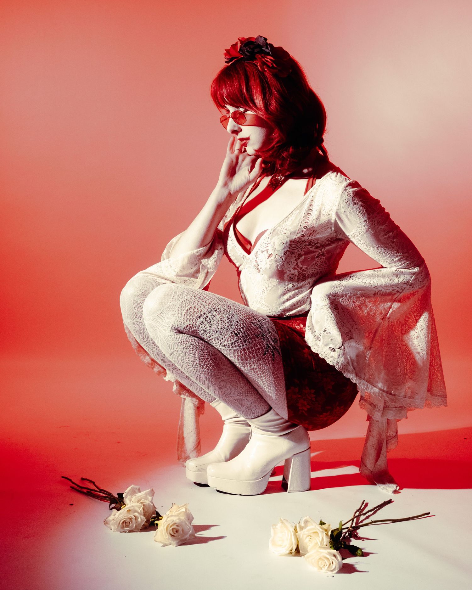 Redhead woman in white lace outfit crouching with roses on red background.