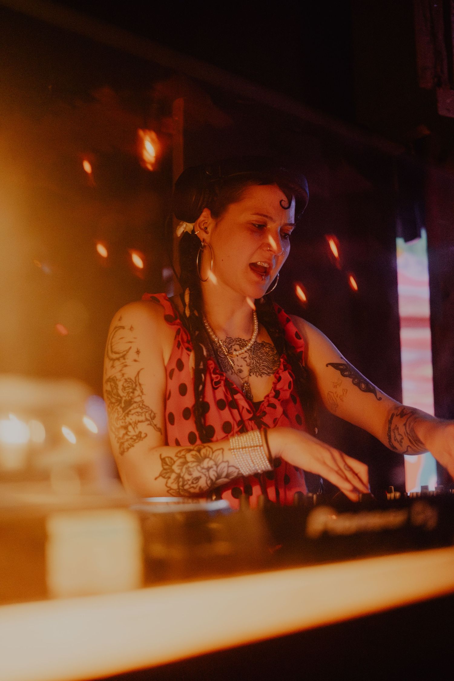Tattooed female DJ in red polka dot dress performing at turntables under warm lights.