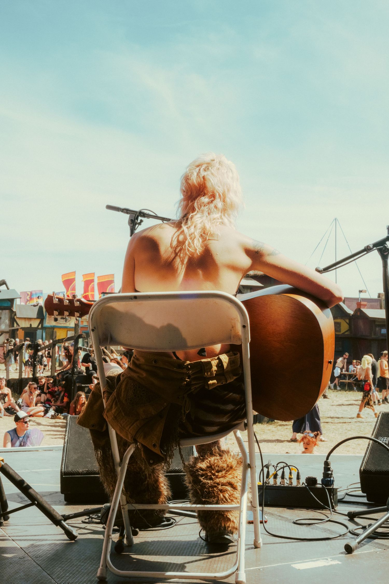 Shirtless blonde musician playing guitar on outdoor festival stage in sunlight.
