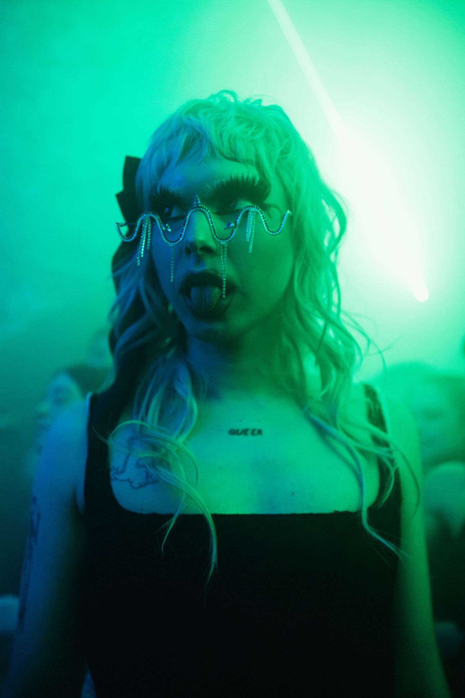 Woman with blonde hair and futuristic spiked glasses poses under vivid green neon lights at a nightclub.