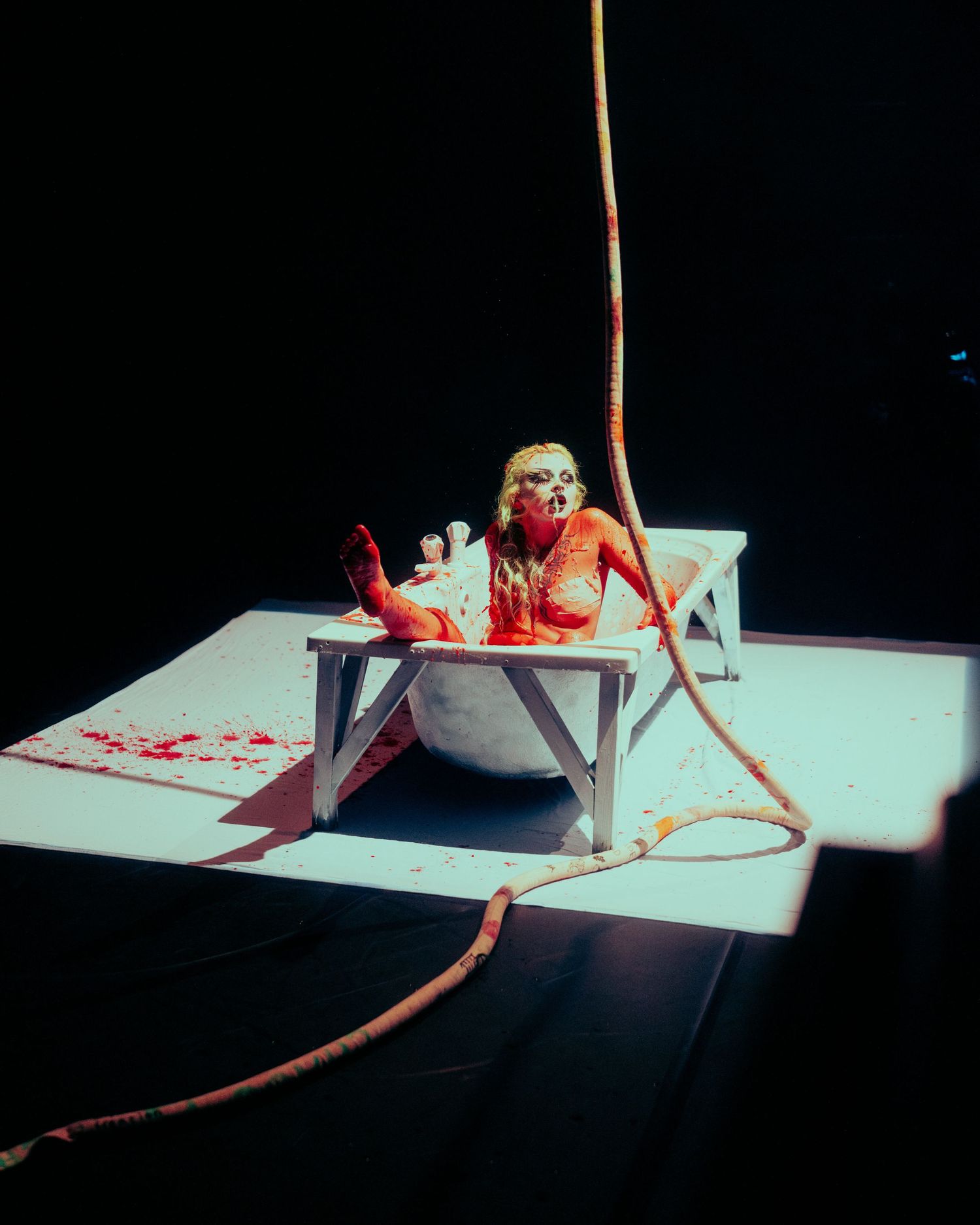 Performer covered in red paint in bathtub during dramatic dark stage production.