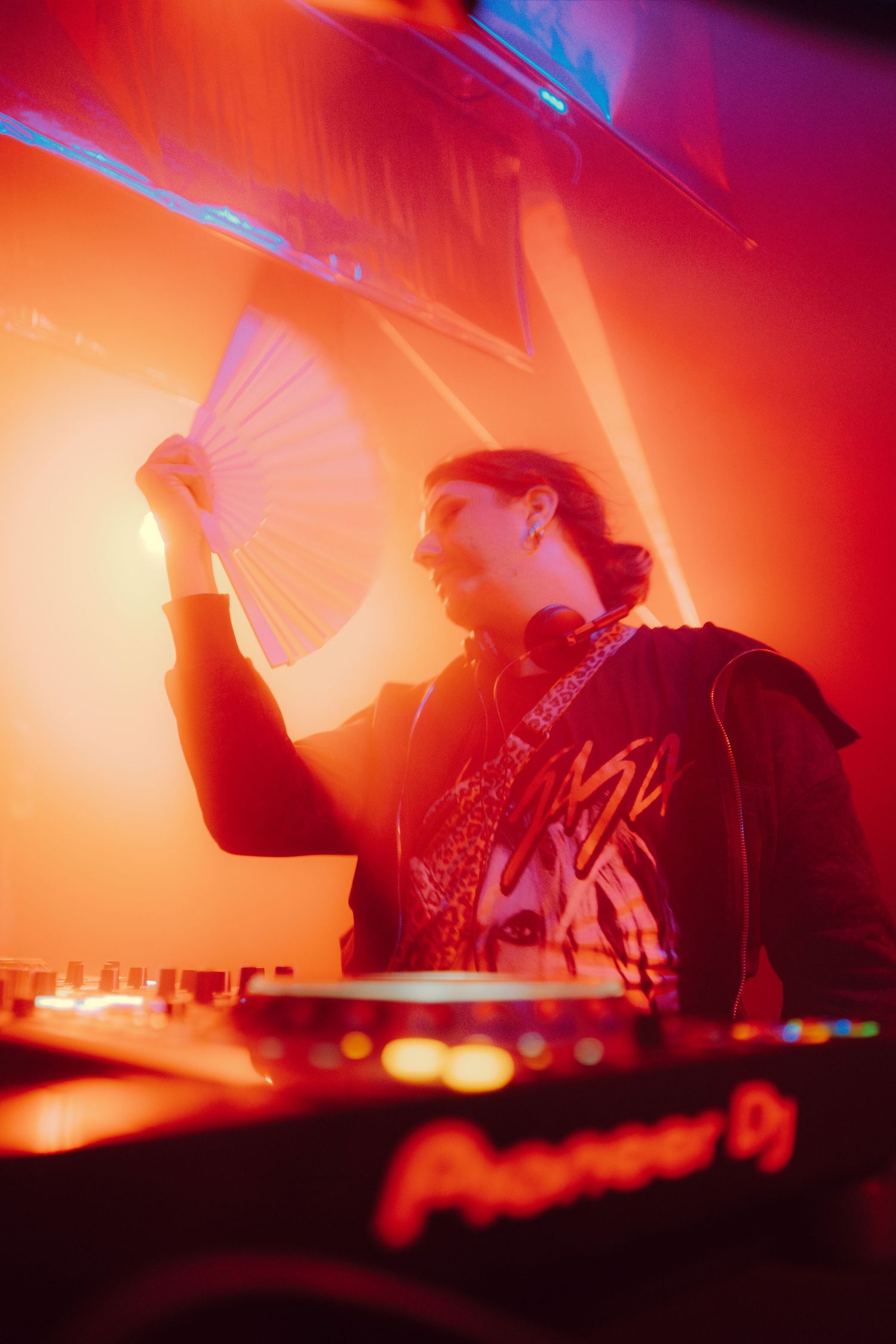 DJ holding a white hand fan while mixing on Pioneer DJ decks under vibrant orange stage lighting.