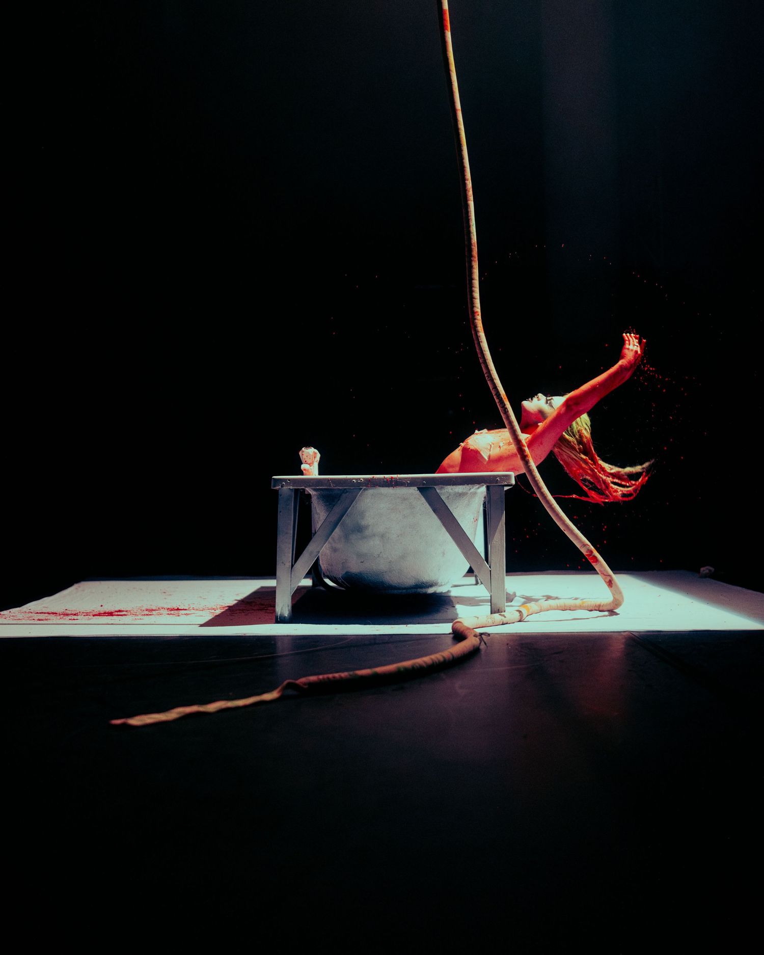 Acrobat in red costume balances on a rope beside a white bathtub prop on a dimly lit stage.