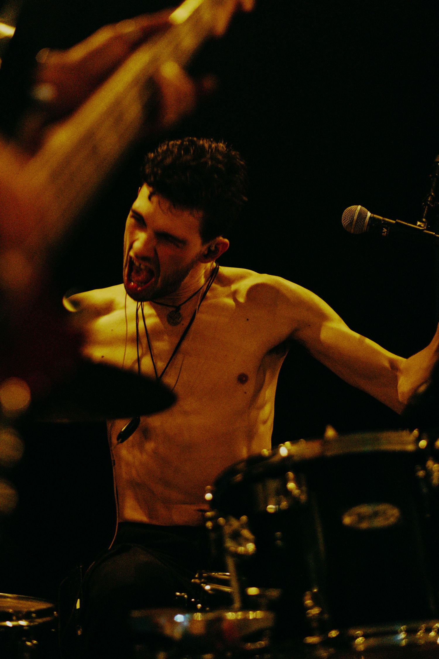 Shirtless male drummer playing intensely on stage under warm golden stage lighting.