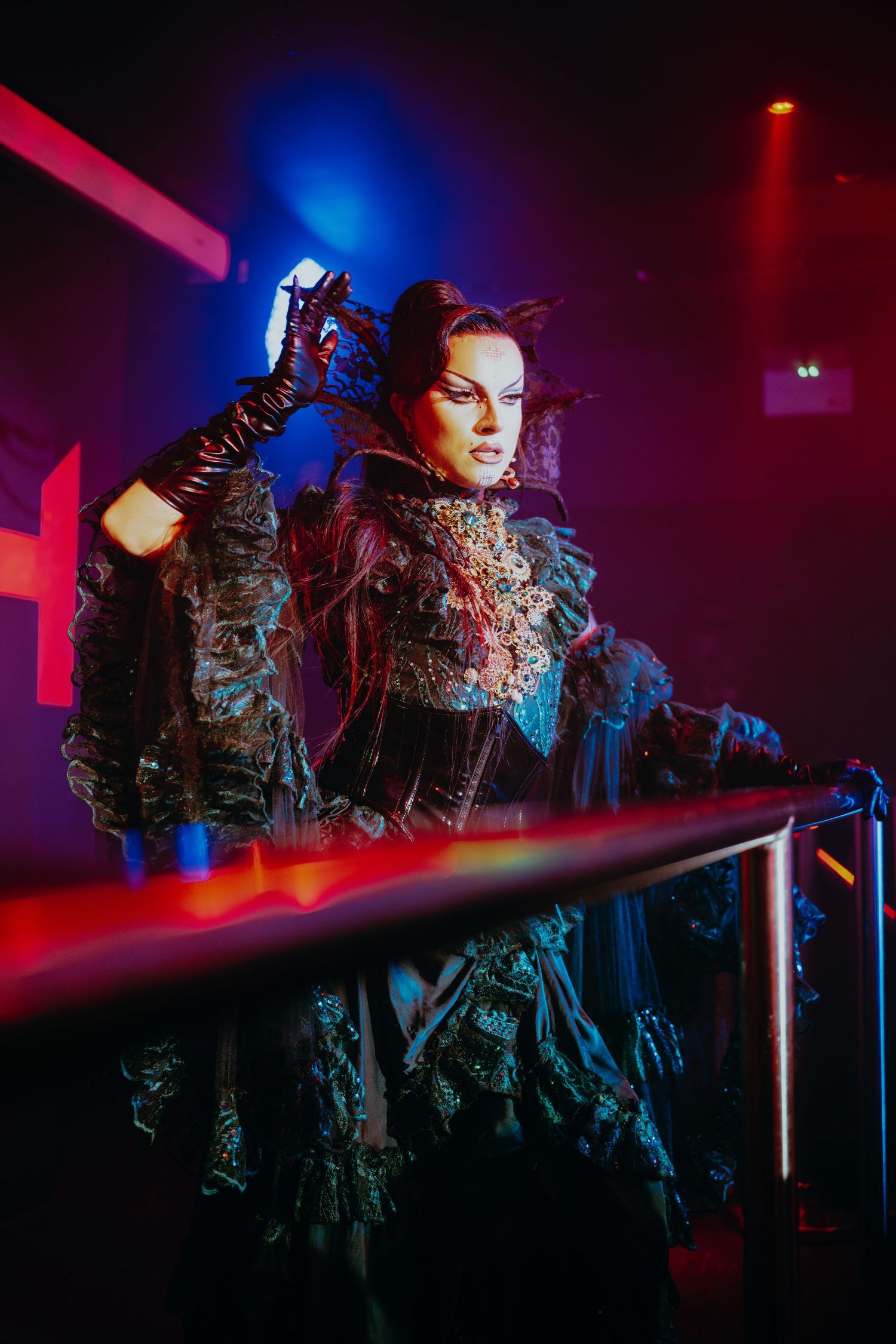 Drag performer in elaborate dark feathered costume poses under purple and red club lights.