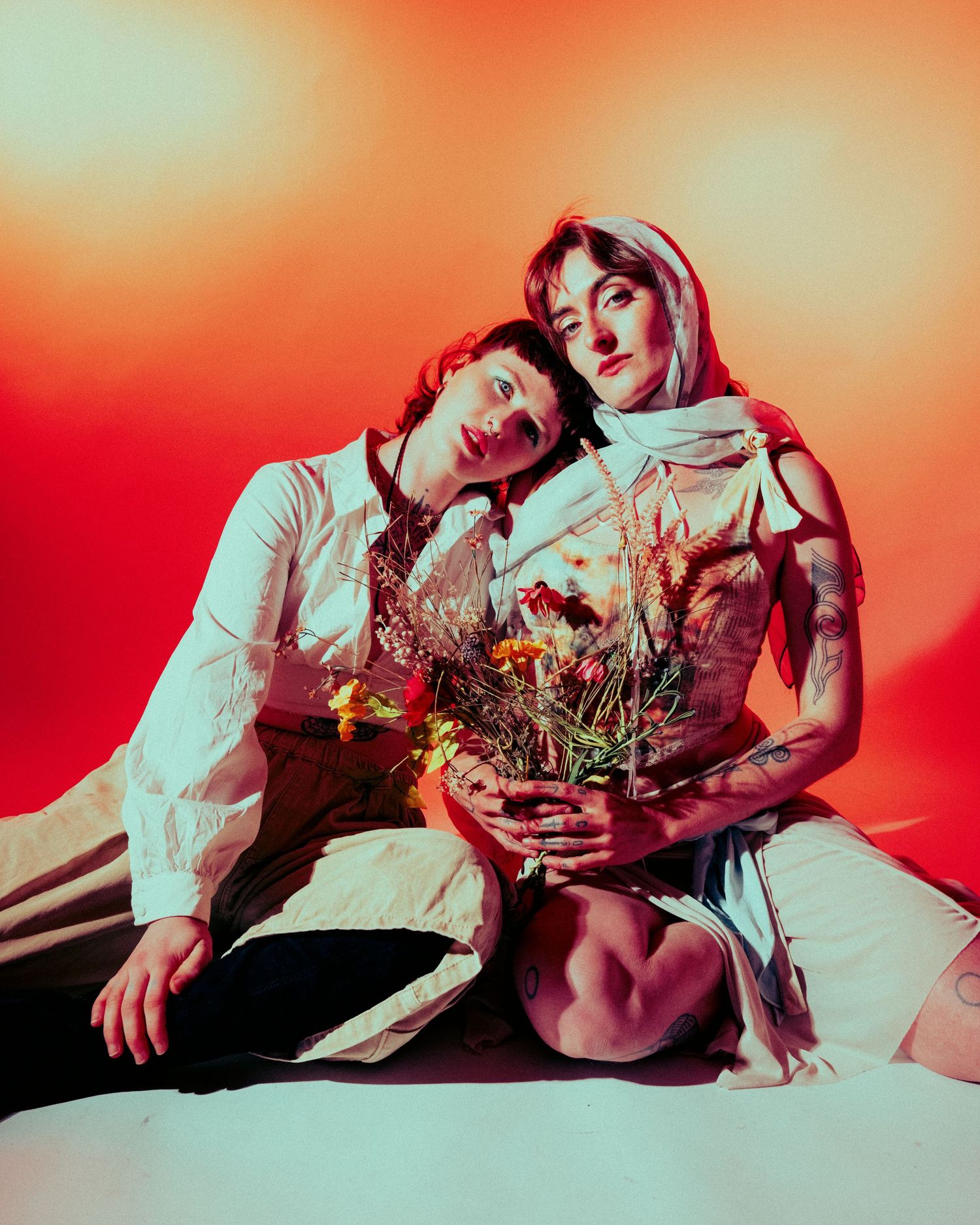 Two people sitting together holding wildflowers against vibrant orange-red backdrop.