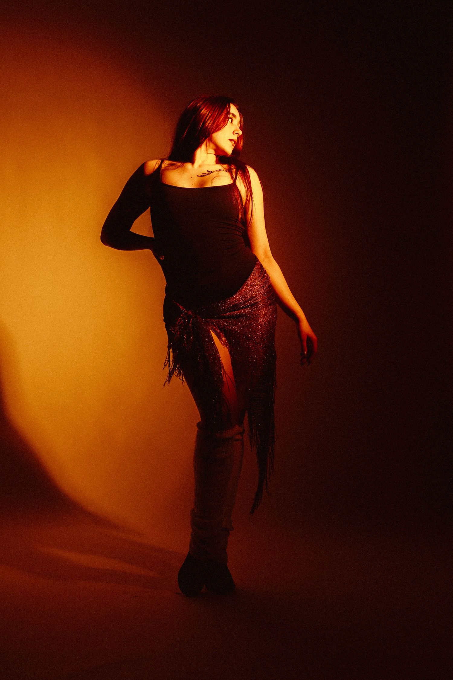 Woman in black top and fringe skirt poses under warm orange studio lighting.