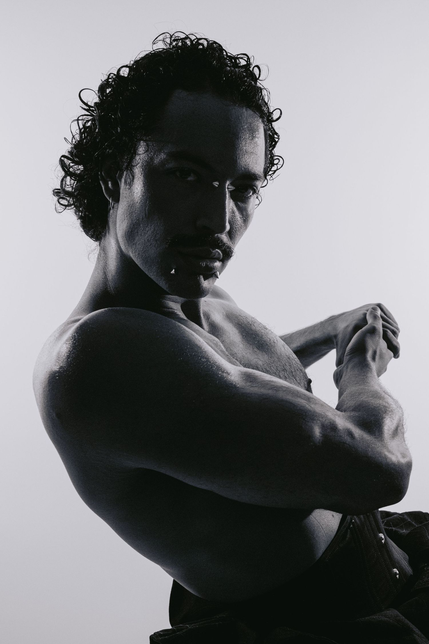 Muscular shirtless man with curly hair poses in dramatic black and white portrait.
