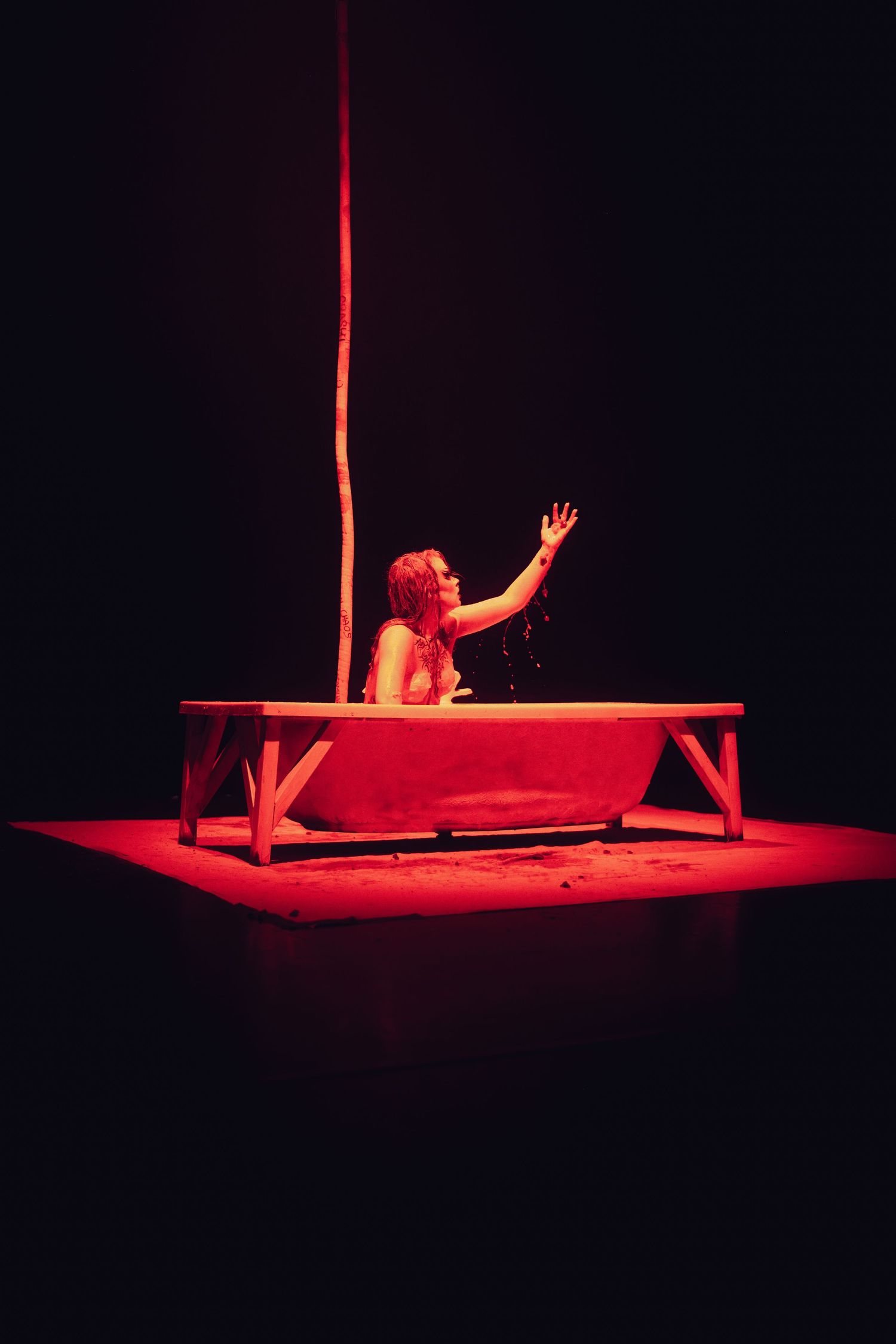Performer in red lighting reaches upward from a white bathtub during an atmospheric theatrical stage production.