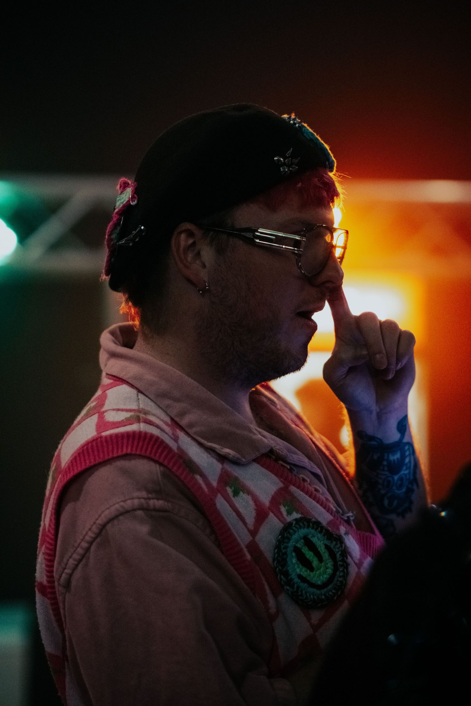 Man in pink jacket and cap touching his nose under colorful concert lighting.