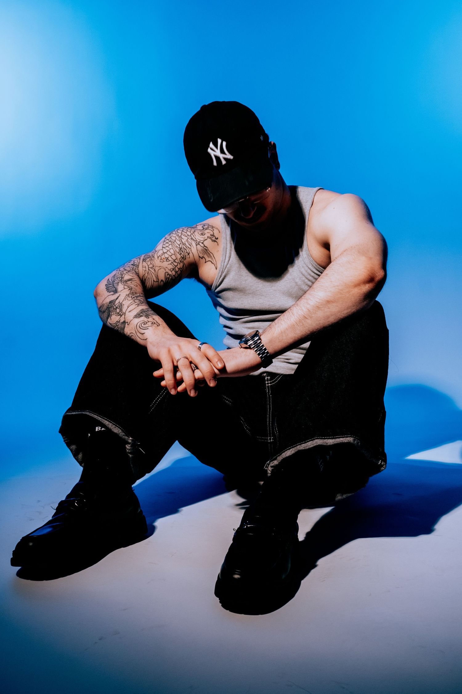 Tattooed man in NY cap and tank top sitting on floor under blue light.