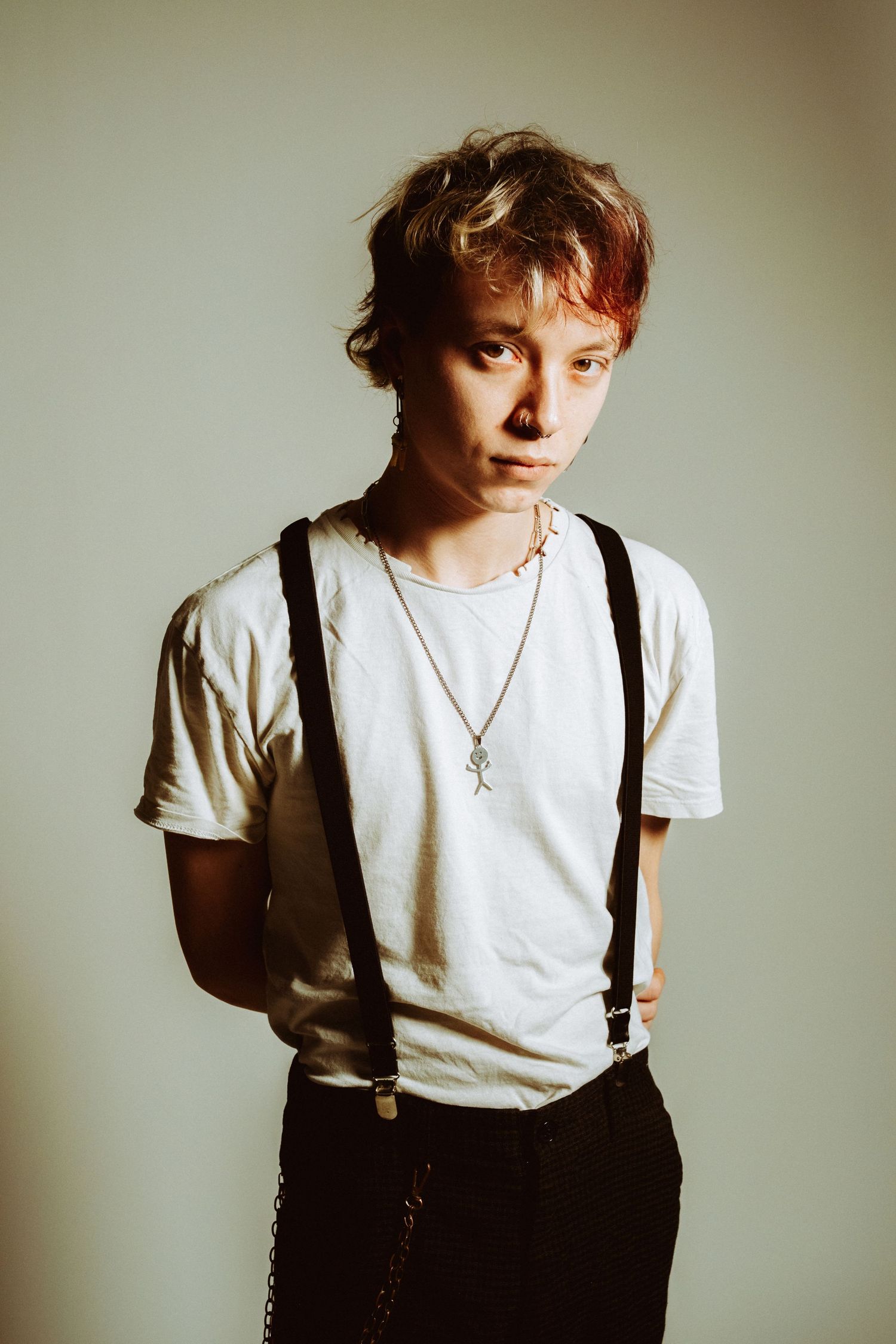 Young person with reddish hair wearing white tee and black suspenders in studio.