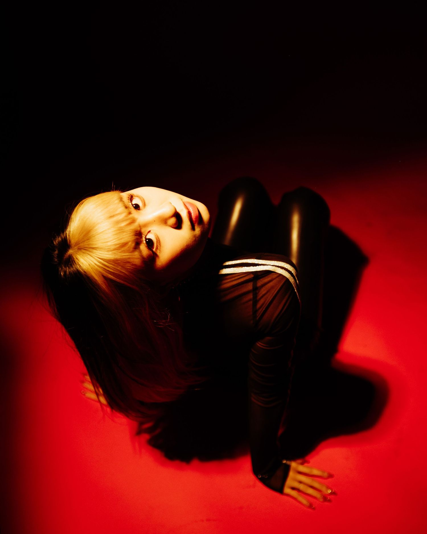 Person in black leather outfit posed on red floor under dramatic lighting.