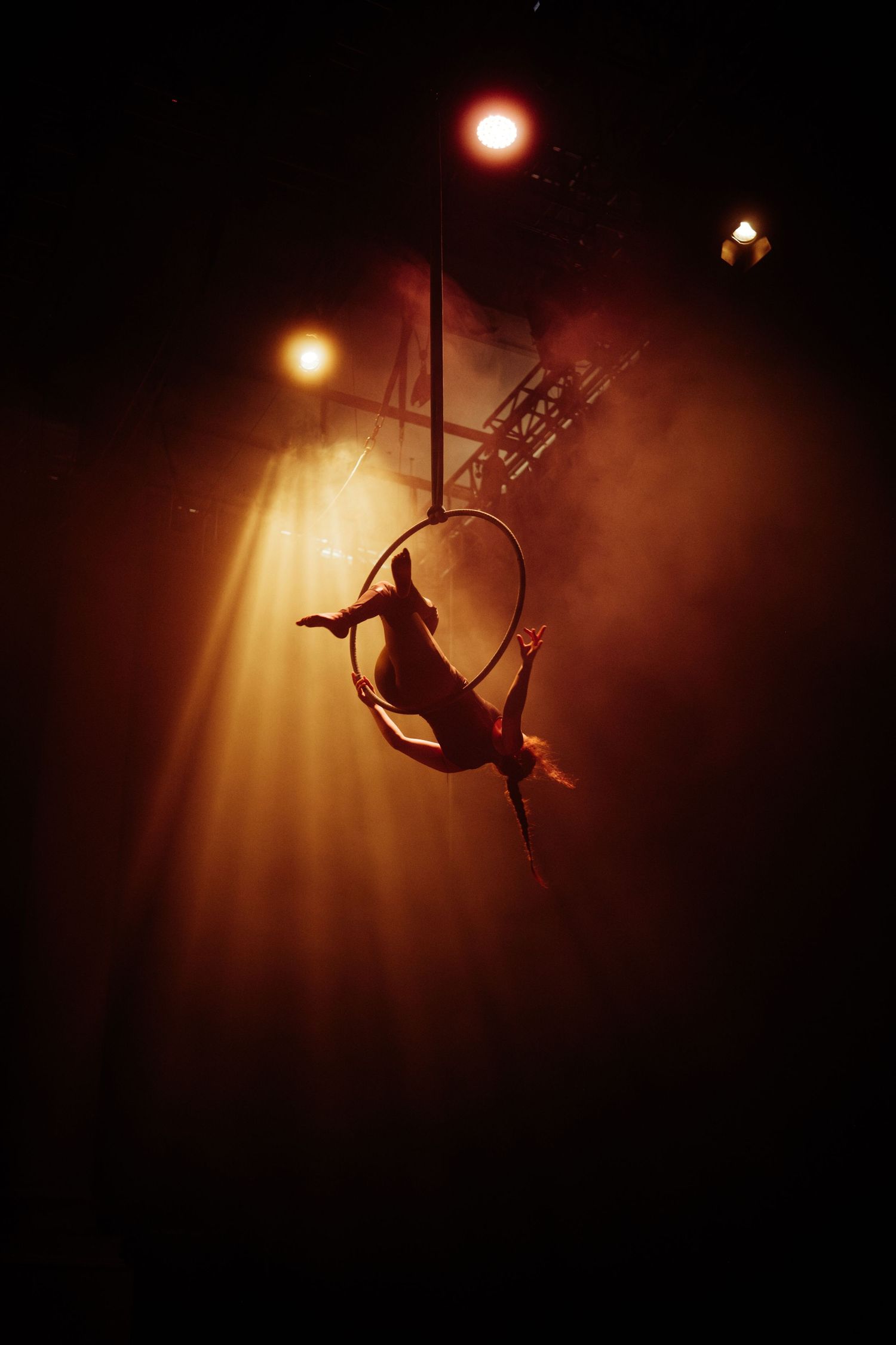 Aerial acrobat performing silhouetted in a hoop under dramatic warm stage lighting.