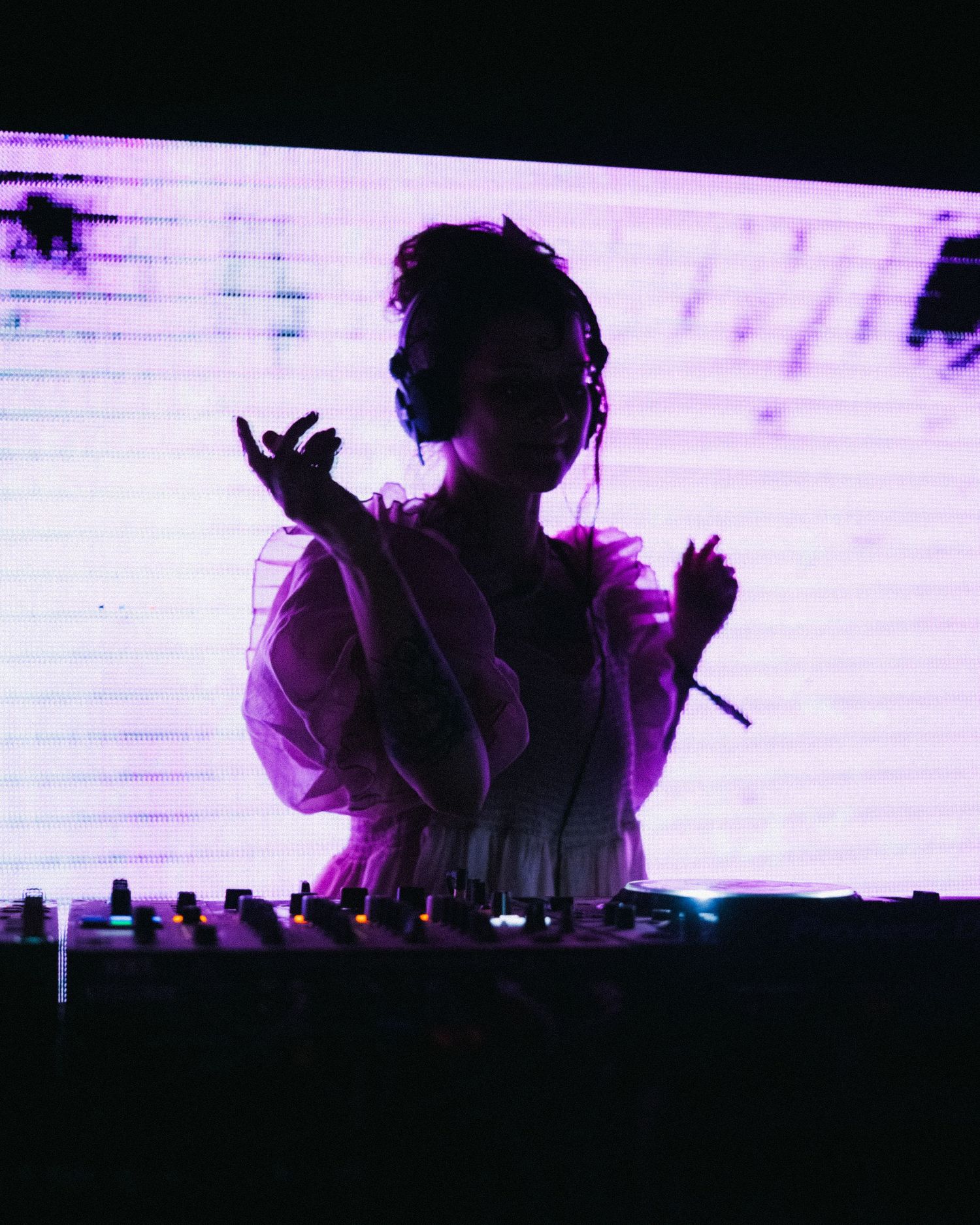 Silhouetted DJ in a ruffled white outfit performs at a mixing board against a glowing purple screen.