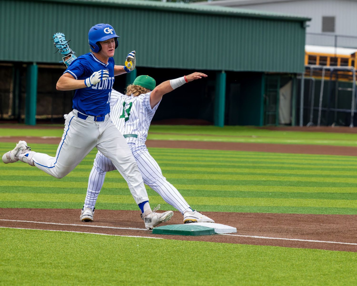 Baseball player in blue uniform rounds first base as green and white fielder attempts to make the out.