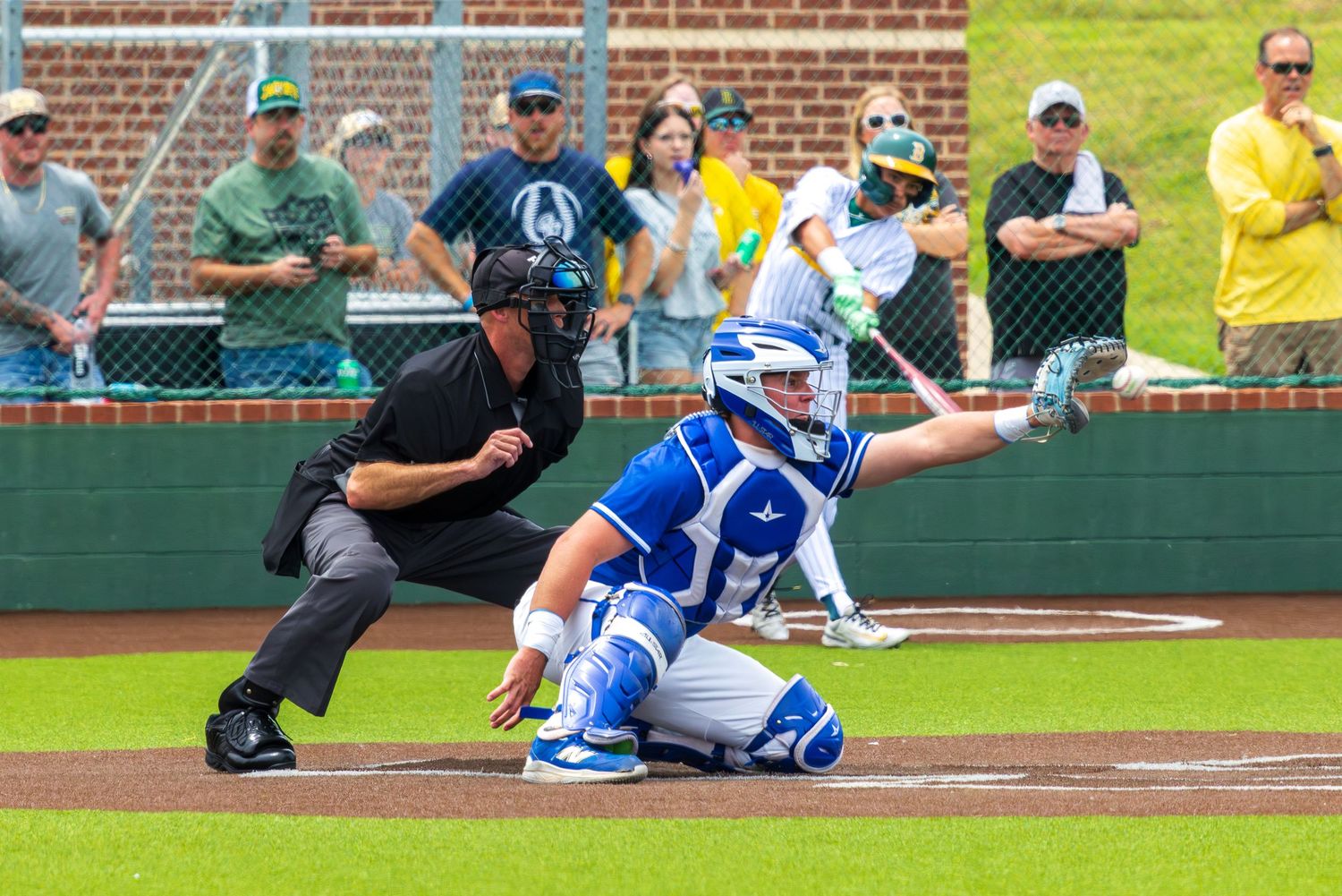Baseball catcher in blue gear crouches at home plate as umpire watches the pitch during a sunny outdoor game.