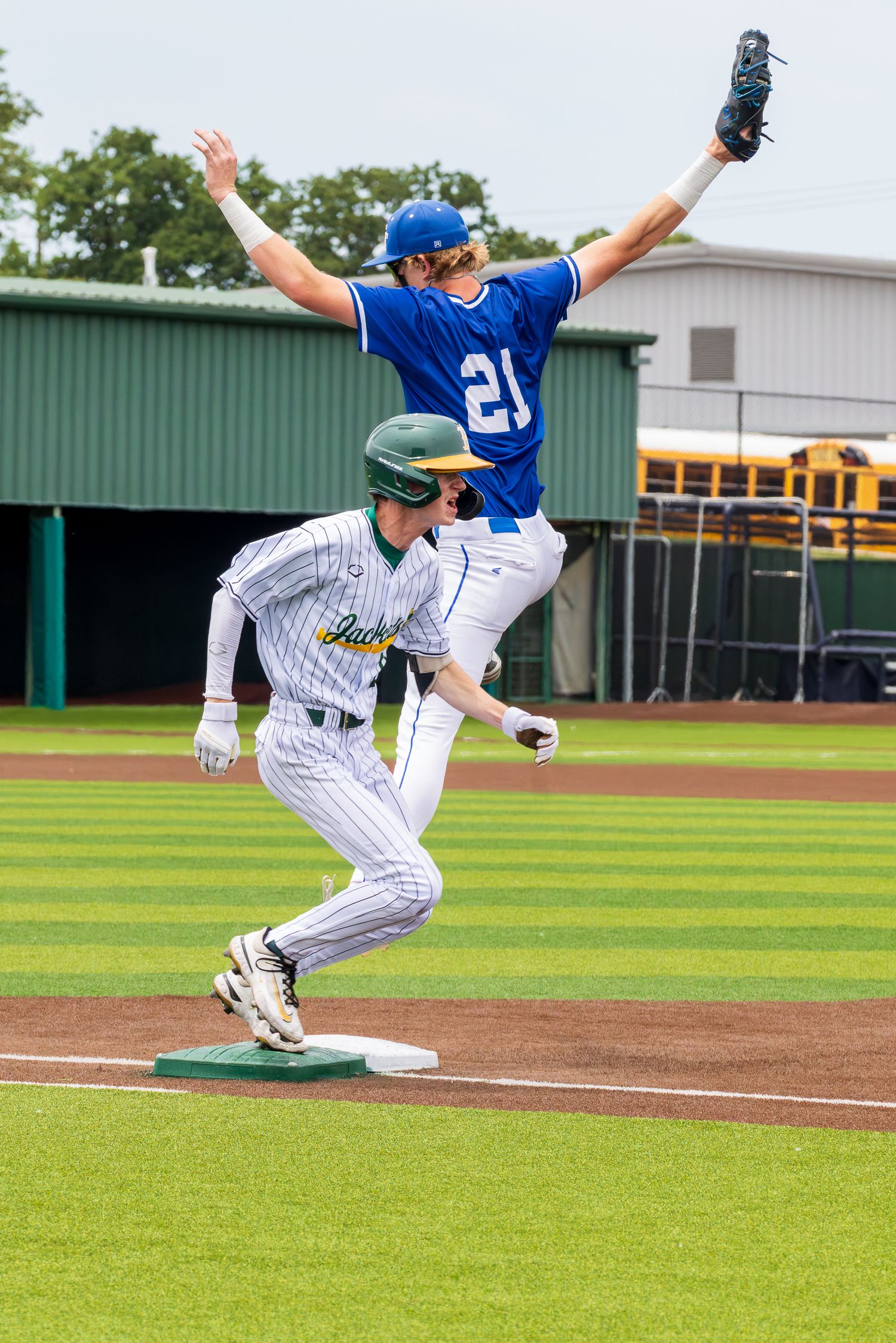 Baseball player in green helmet rounds base as player #21 in blue leaps for high throw during youth baseball game.
