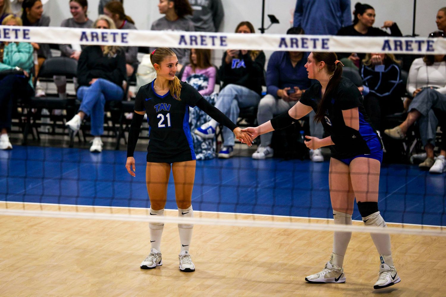 Two female volleyball players in blue uniforms exchange a fist bump near the net during a match.